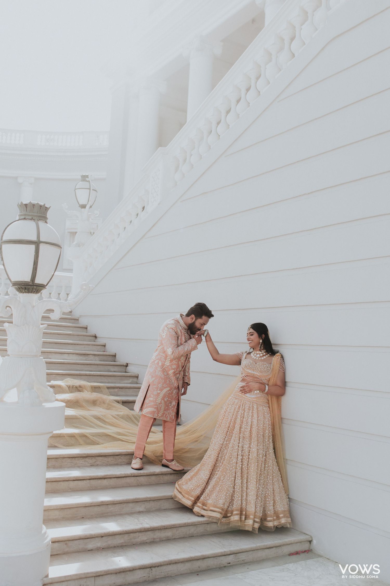 Couple in blush pink Indian wedding attire pose on white marble stairs, groom kissing bride's hand.