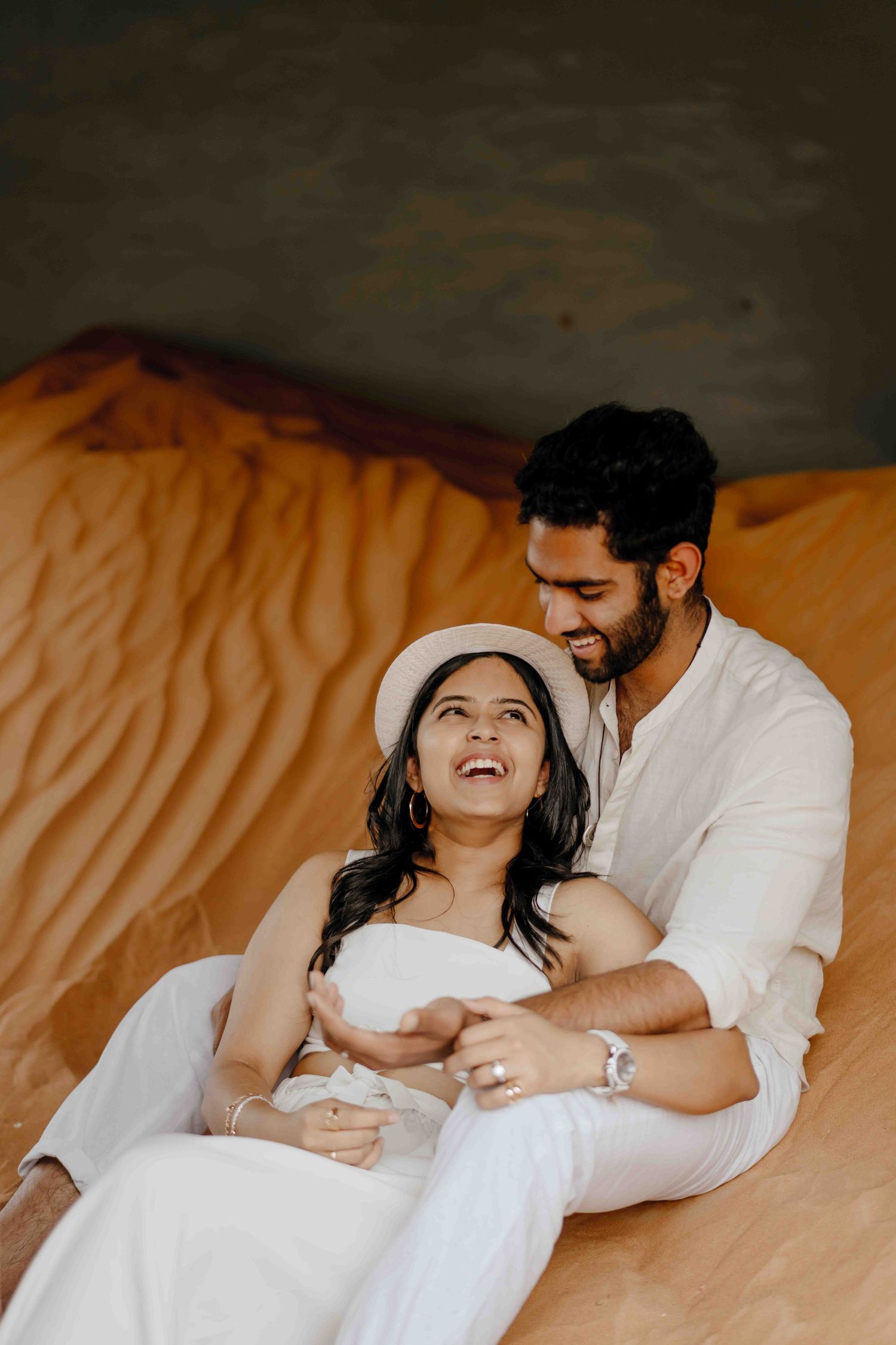 Happy couple in white outfits sitting together on golden sand dunes, laughing and enjoying a romantic desert photoshoot.