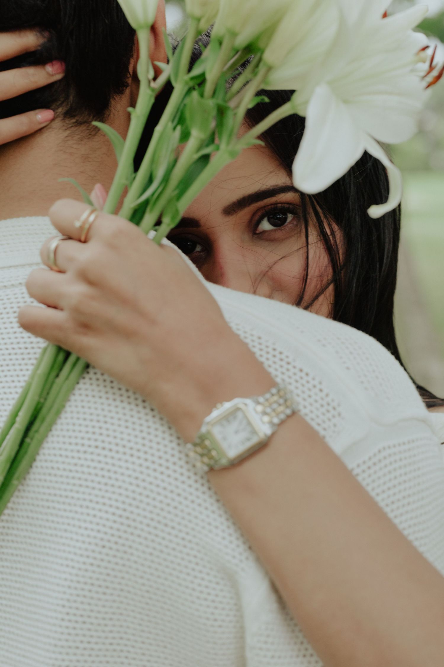 Woman with dark hair peeks over a bouquet of white lilies while embracing someone, wearing a silver watch and rings.