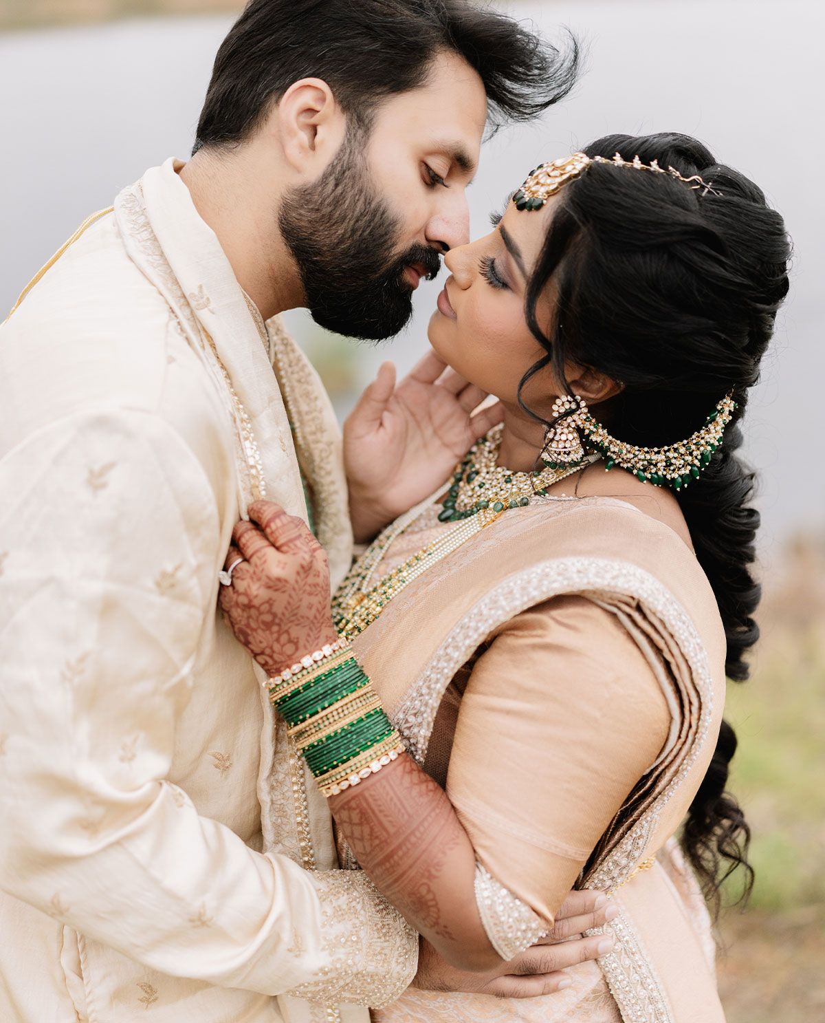 Indian bride and groom in traditional attire share an intimate romantic moment outdoors near a lake.