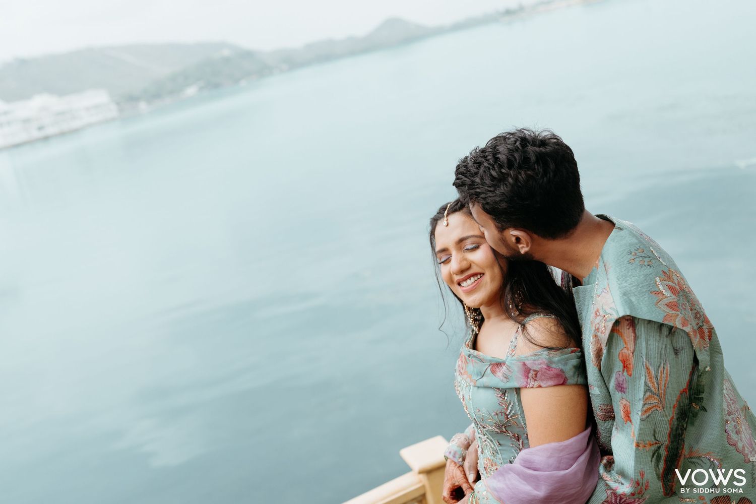 Romantic couple sharing a tender moment by a serene lake, woman smiling joyfully in pastel outfit at a scenic waterside location.
