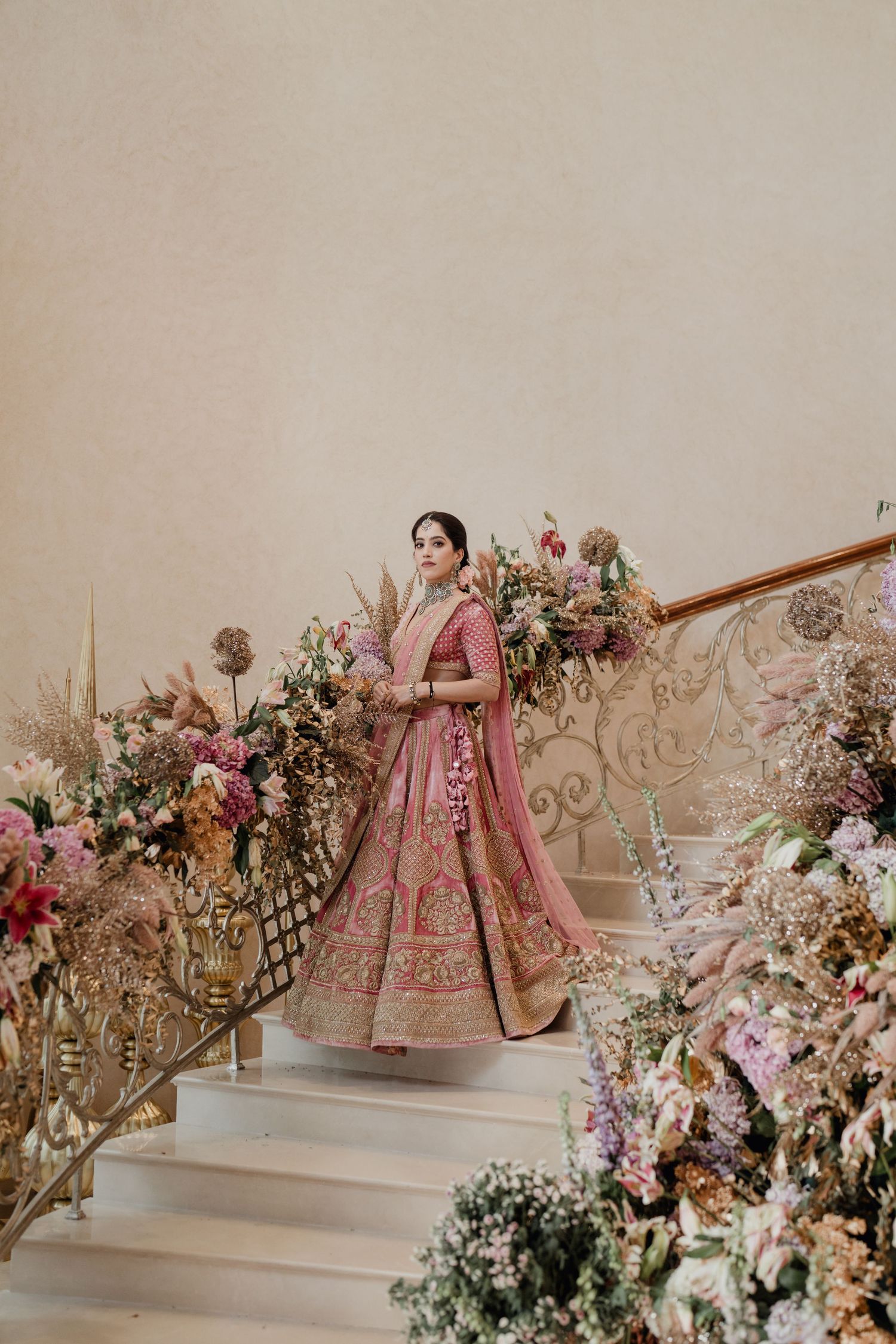 Bride in pink lehenga standing on floral-decorated staircase in elegant bridal portrait.