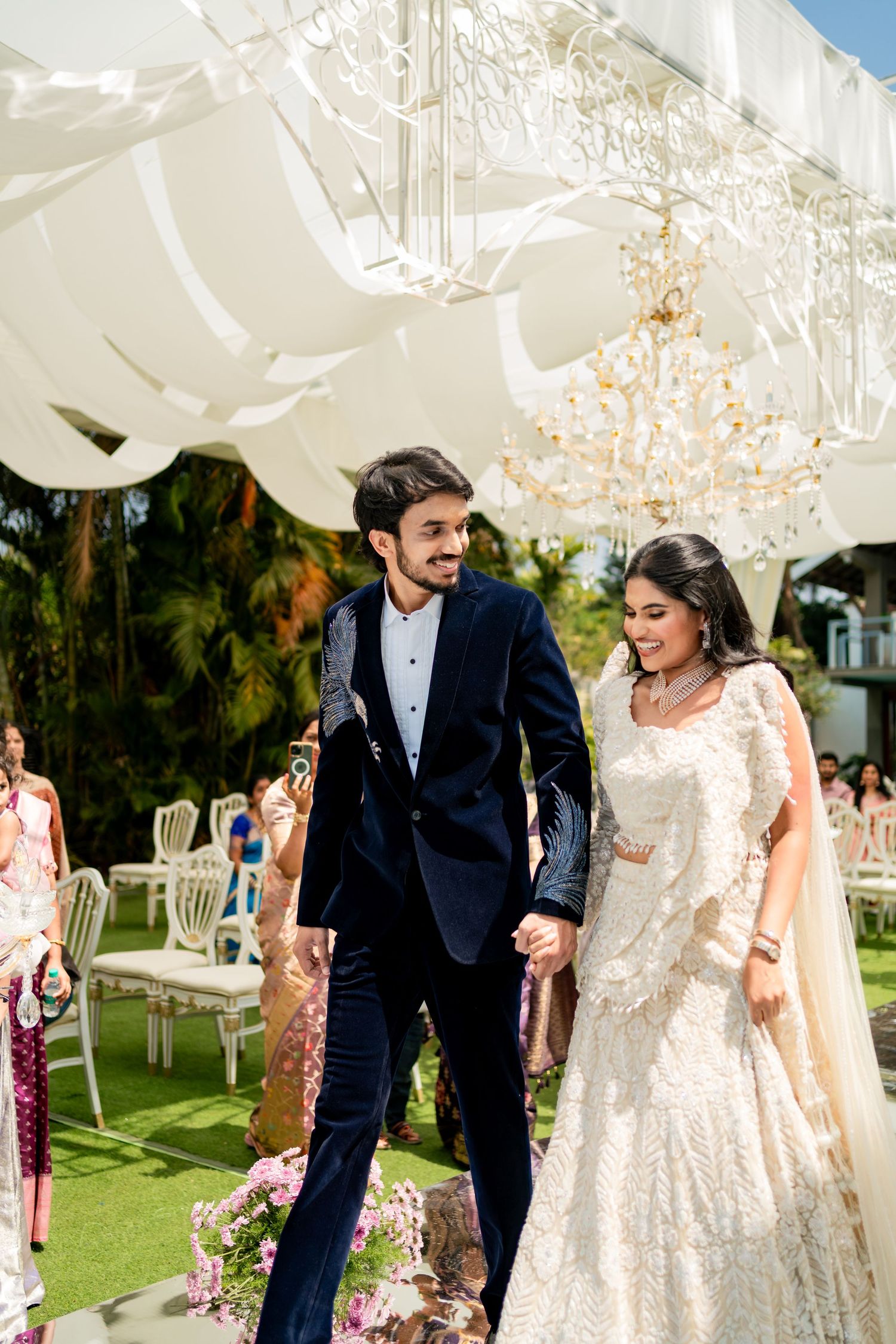 Indian couple in elegant wedding attire walking hand in hand under decorative white canopy outdoors.