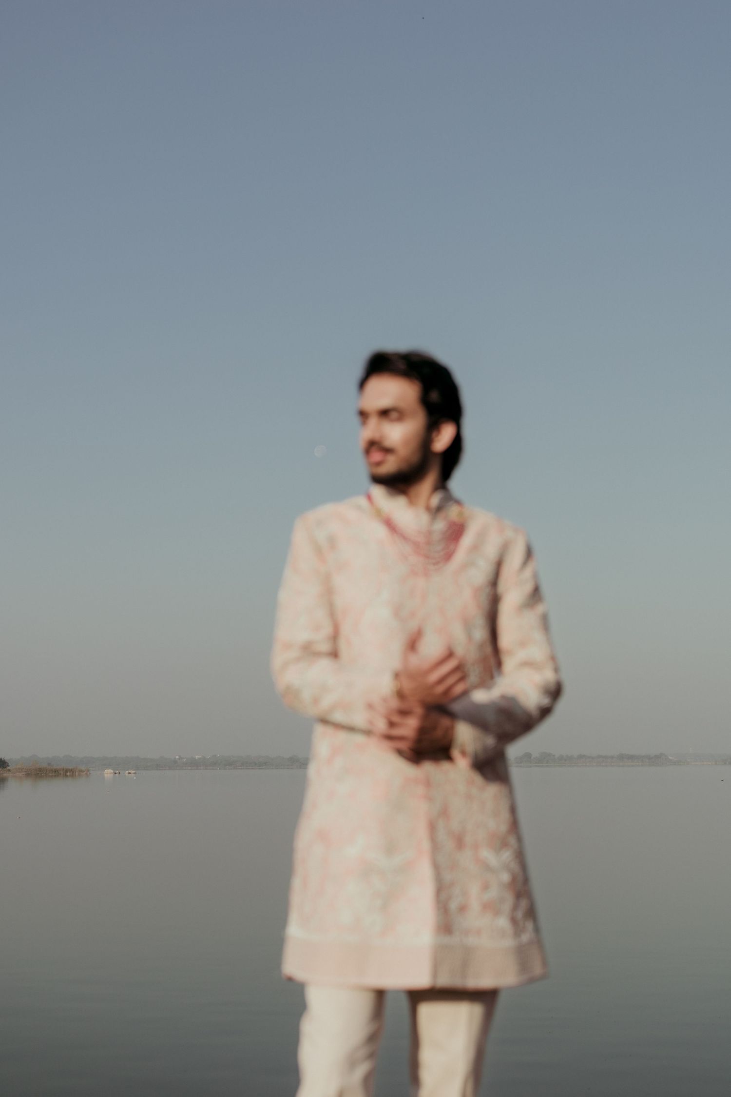Man in embroidered pink sherwani stands by calm water under clear blue sky at dusk.