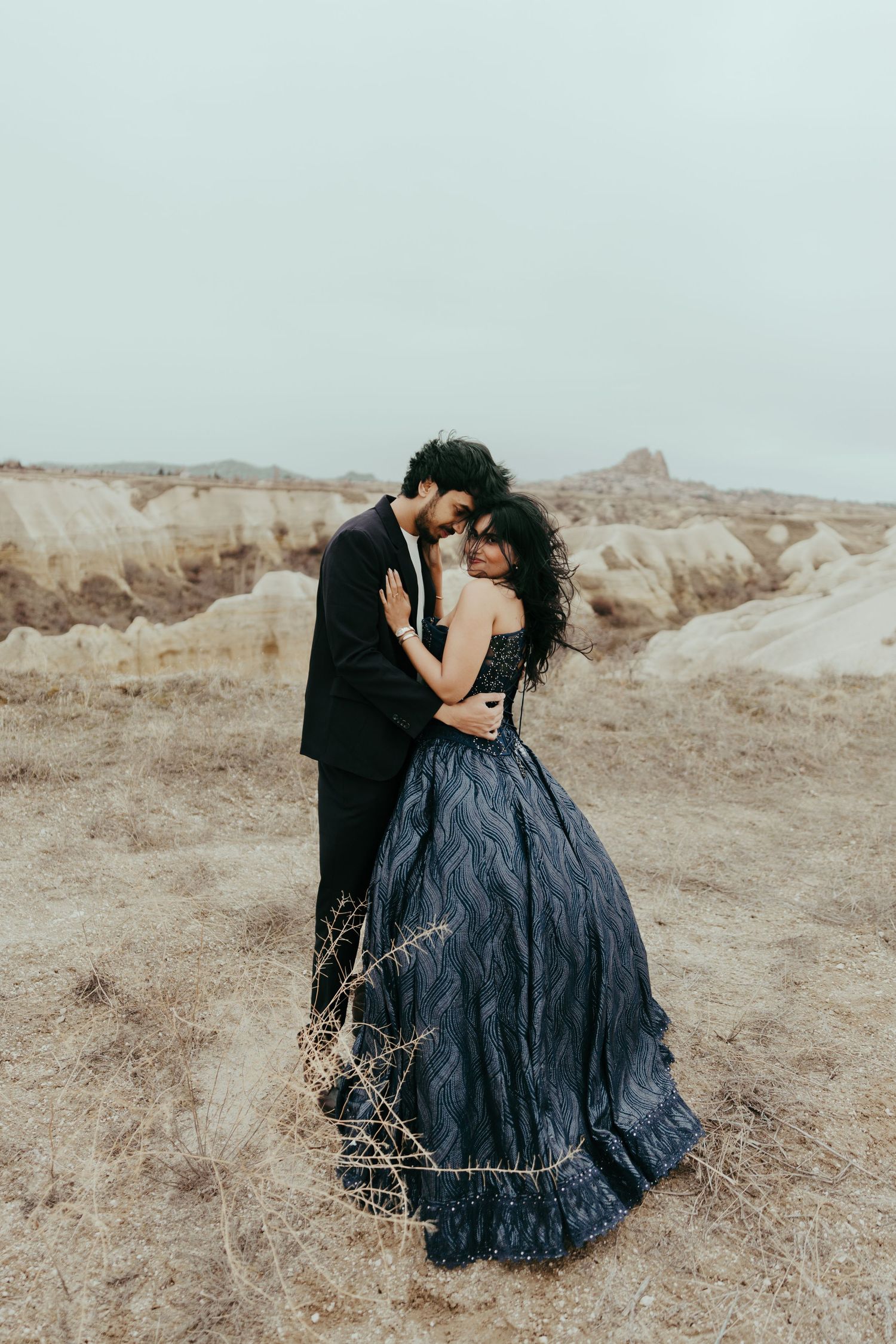 Couple embracing romantically in a dark blue ball gown and black suit in an arid desert landscape.