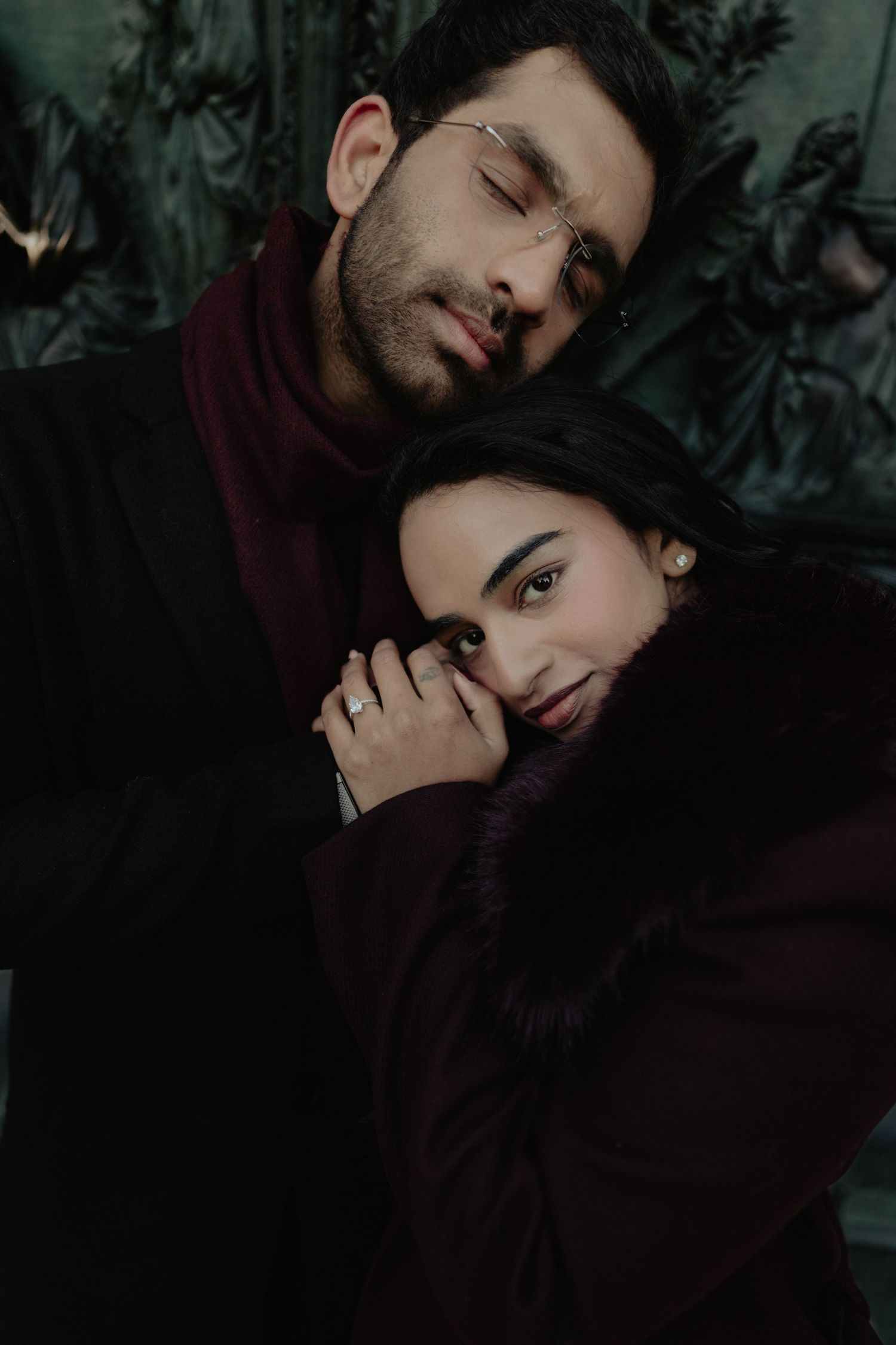 Couple in dark winter coats embracing closely against an ornate bronze sculpture backdrop.