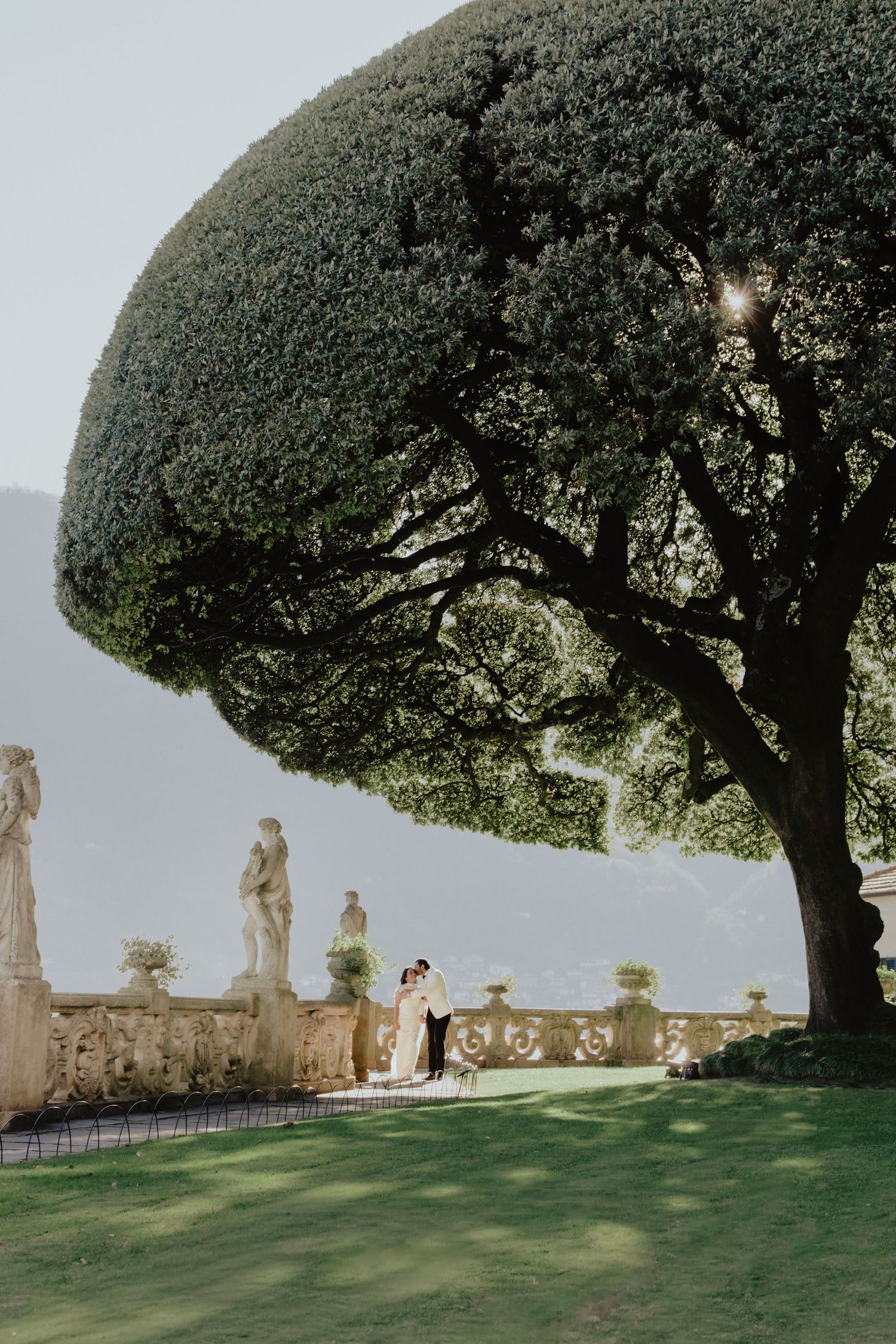 Bride and groom embrace under a majestic topiary tree at an elegant Italian villa garden with stone statues.