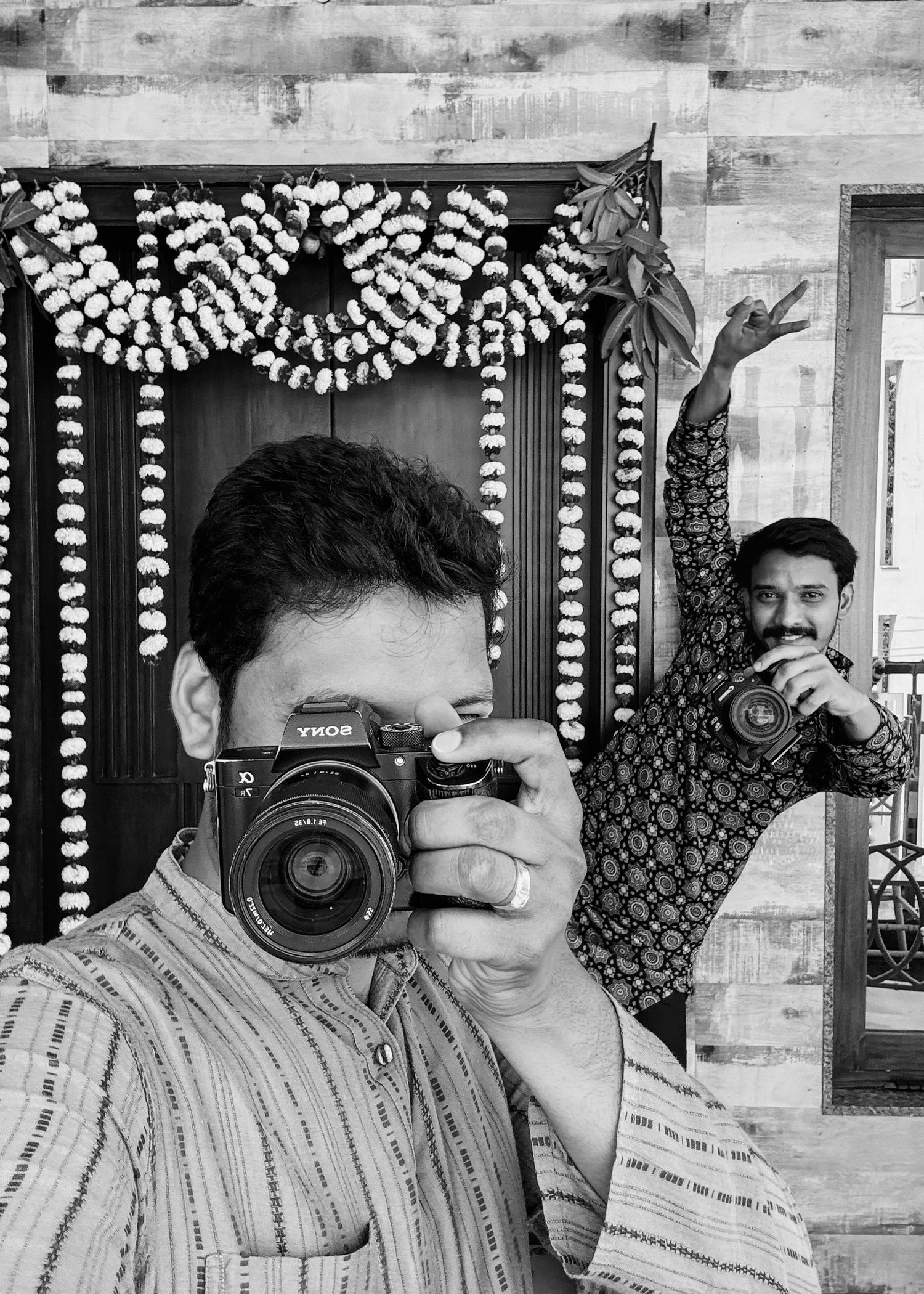 Black and white photo of two men with Sony cameras posing in front of a floral decorated doorway.