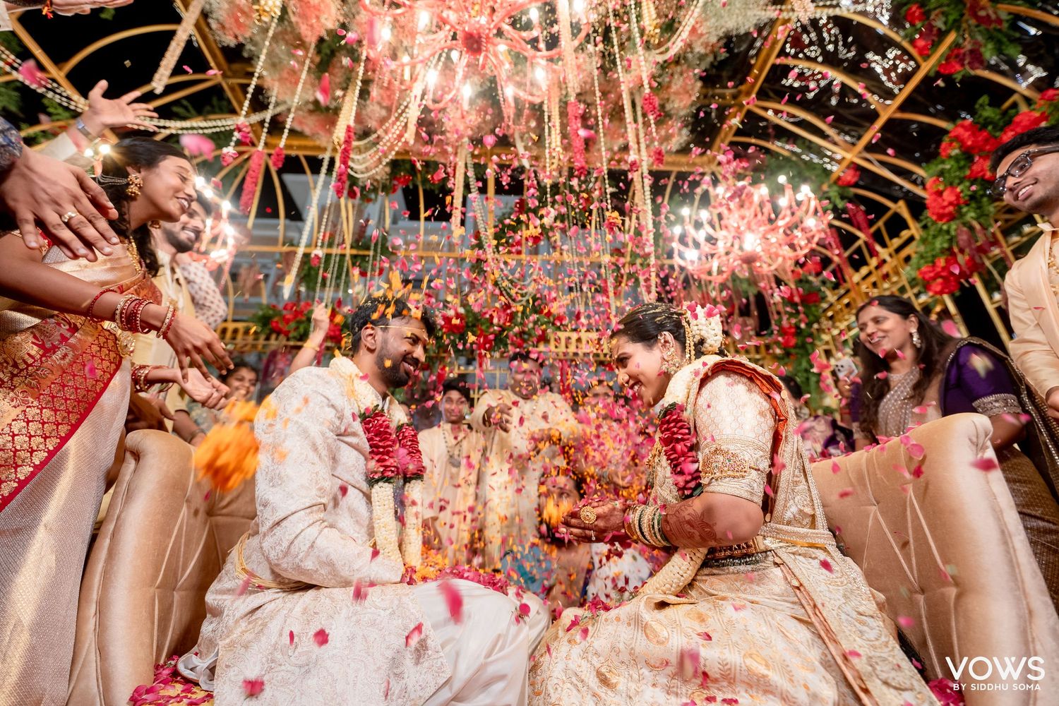 Vibrant Indian wedding ceremony with couple exchanging garlands under floral decor with confetti and guests celebrating.