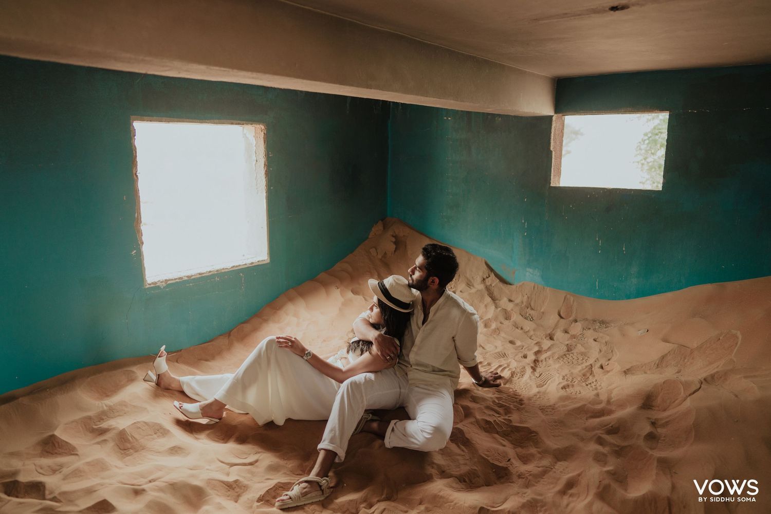 Couple in wedding attire sitting romantically on a sand dune inside a teal-walled room with natural window light.