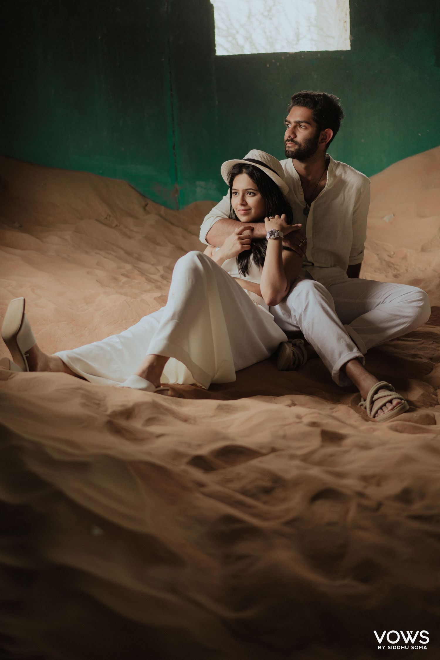 Couple in white outfits sitting on sandy dunes, woman in hat leaning on man, romantic desert pre-wedding shoot.