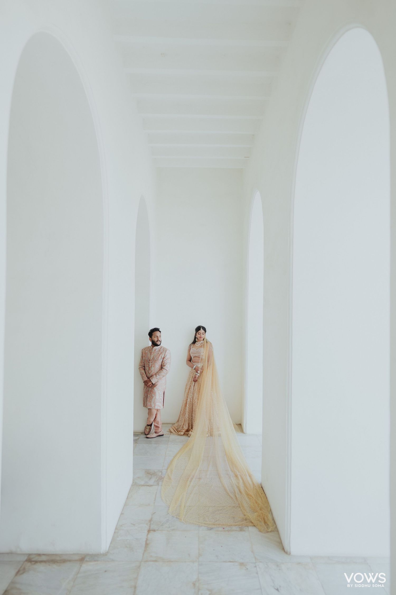 Couple in elegant Indian wedding attire pose in a stunning white arched corridor, bride wearing a flowing golden dupatta.