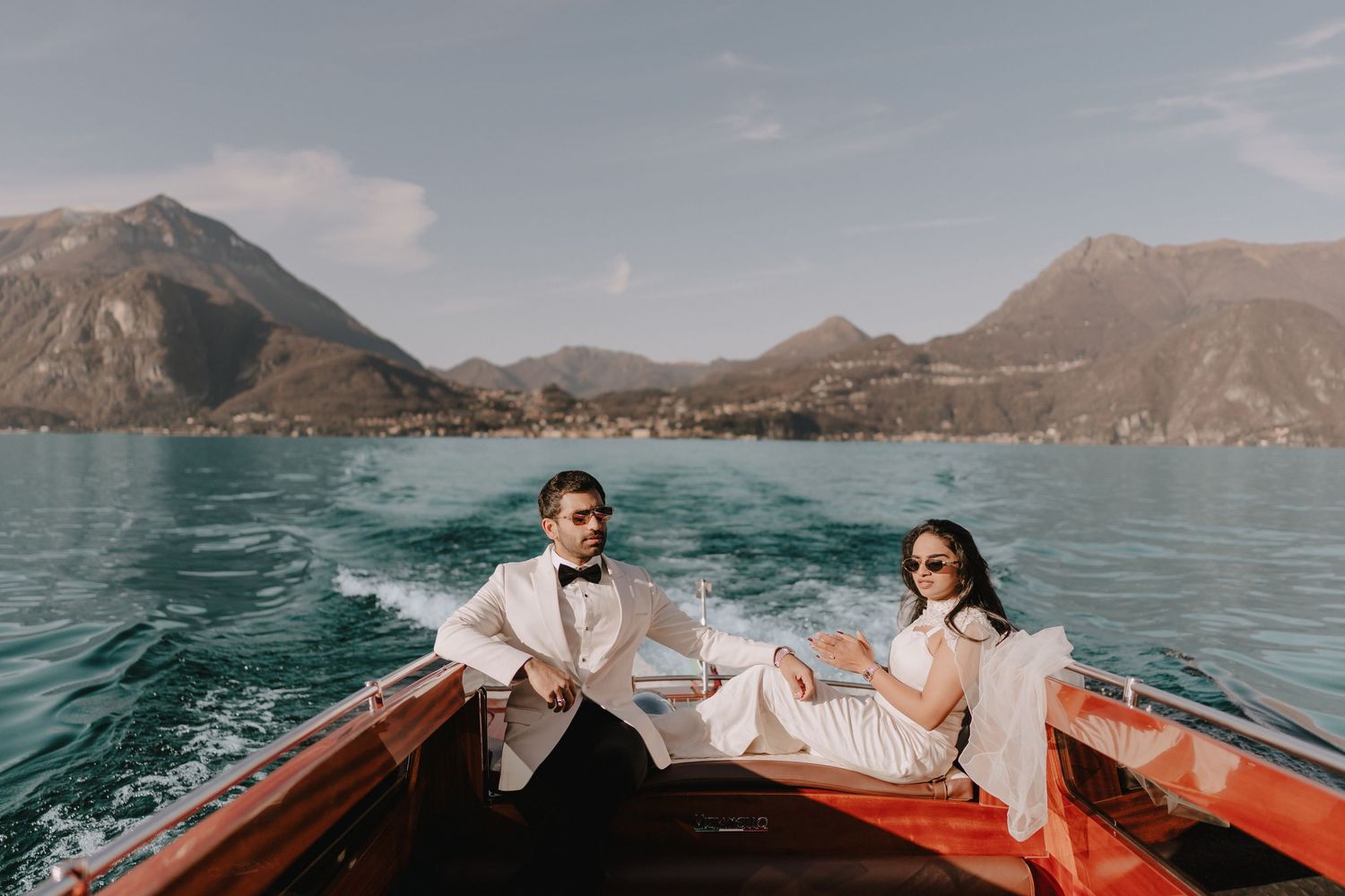 Couple in wedding attire riding a wooden boat on Lake Como with stunning mountain backdrop.
