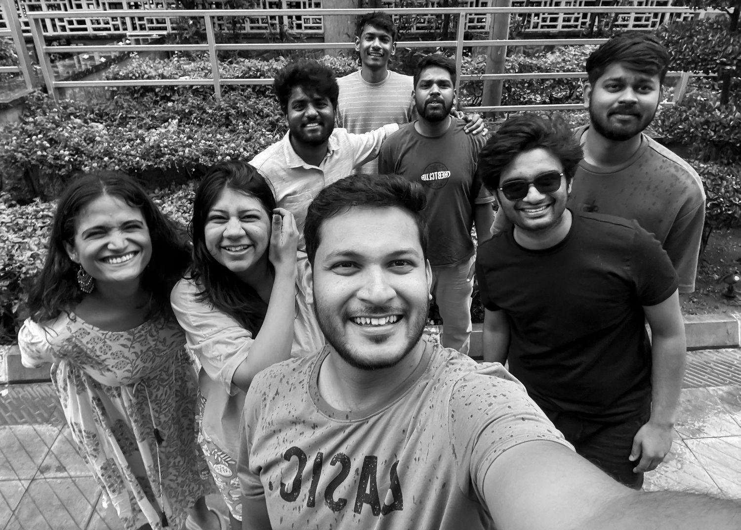 Black and white group selfie of seven smiling friends posing outdoors near greenery and a building.