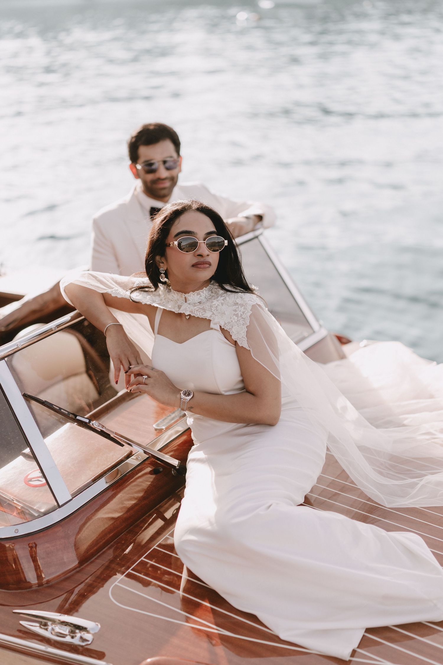 Stylish bride in fitted white gown and sunglasses posing on a luxury wooden boat on the water.