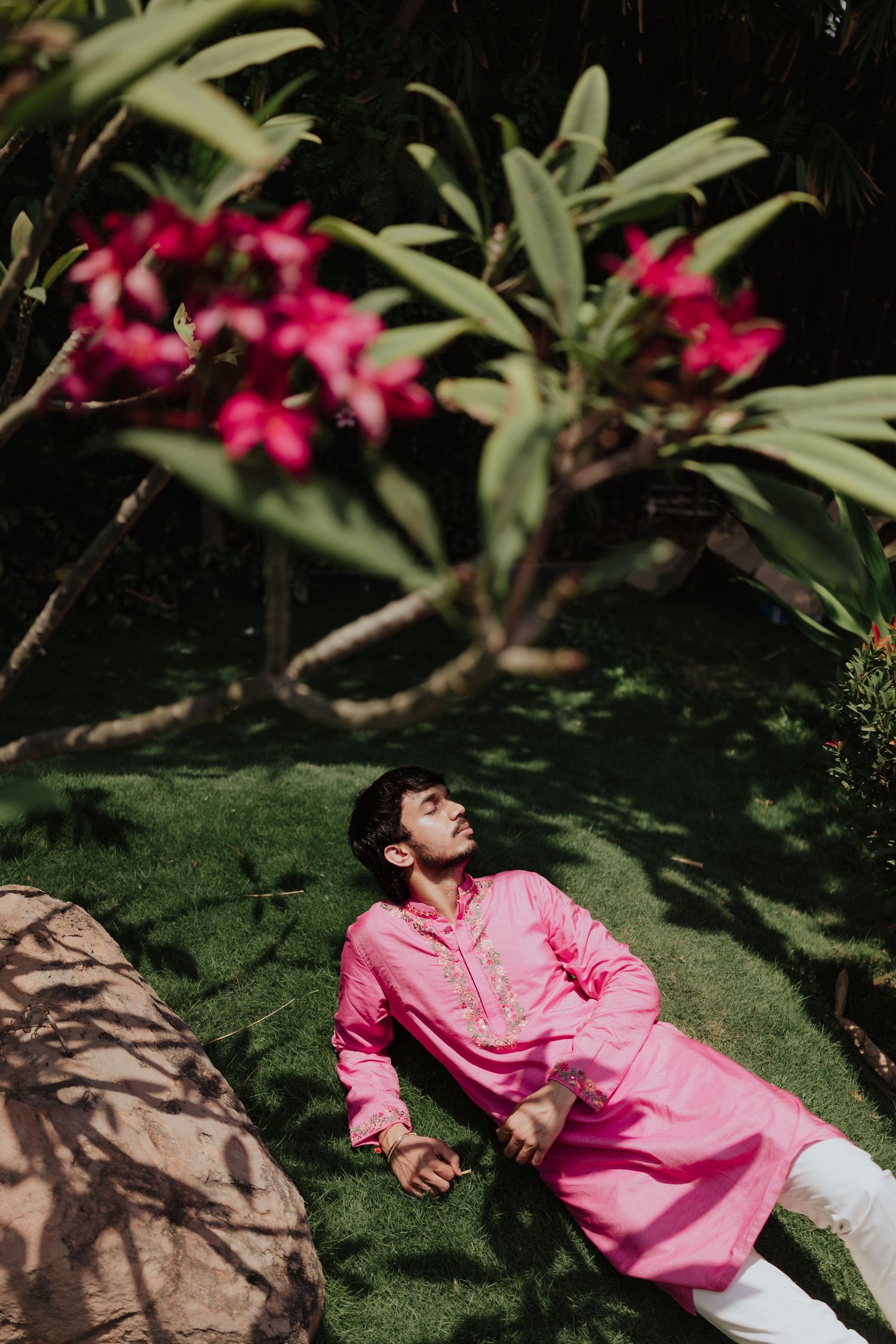 Man in pink kurta lying on grass surrounded by tropical pink flowers in garden.