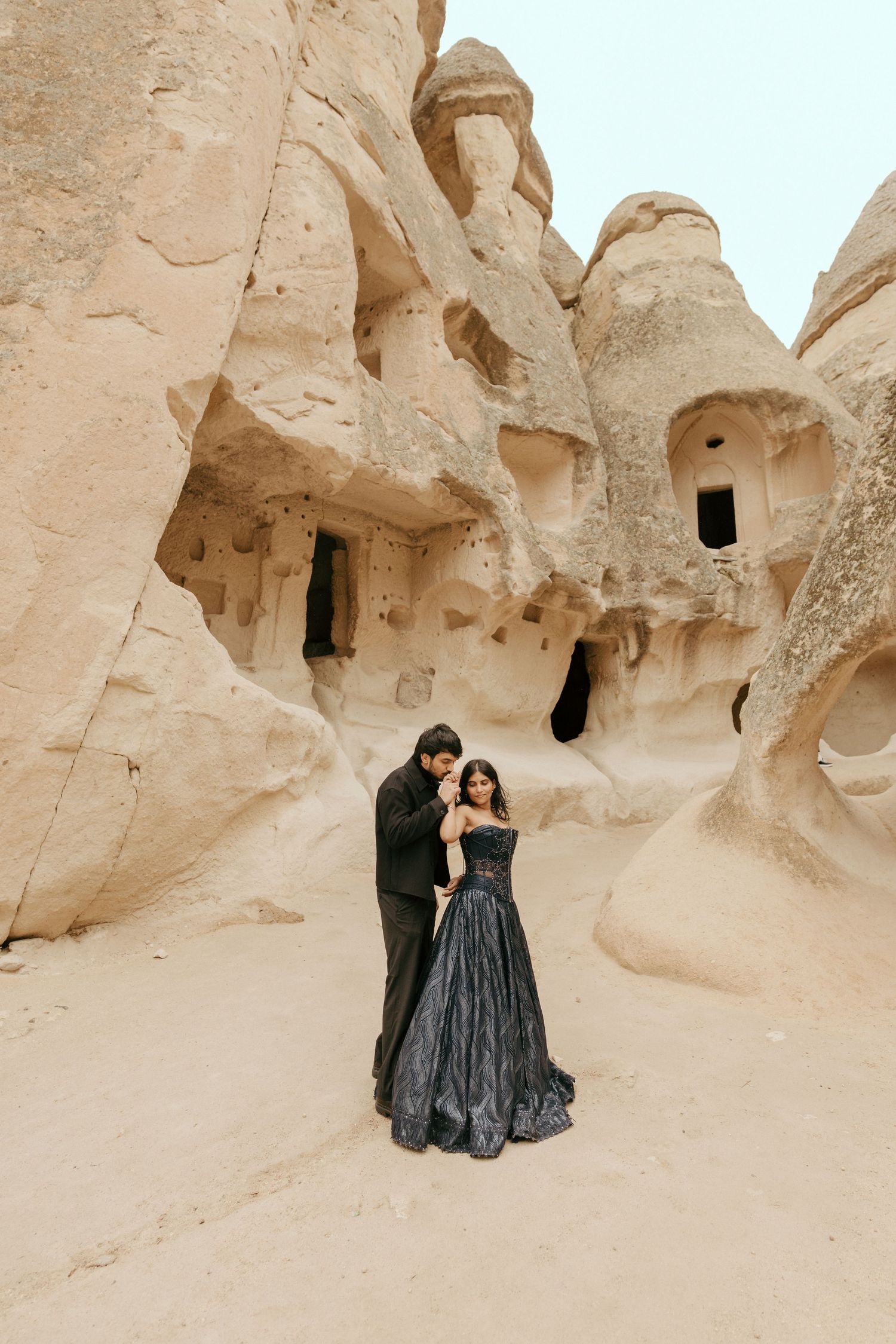 Couple in black formal wear posing before ancient cave rock formations in Cappadocia, Turkey.