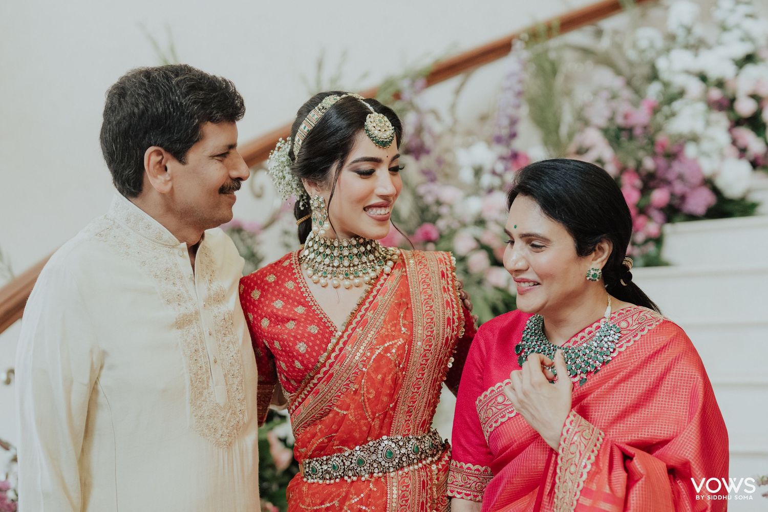 Bride in red saree with gold jewelry smiles with family at Indian wedding ceremony.