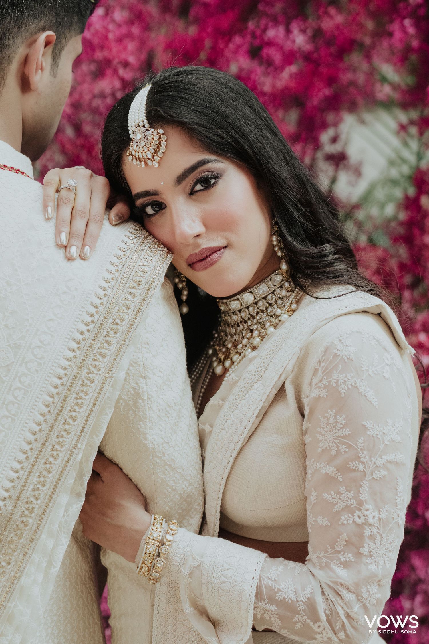 Bride in ivory lehenga with kundan jewelry leans on groom, pink floral backdrop, traditional Indian wedding portrait.