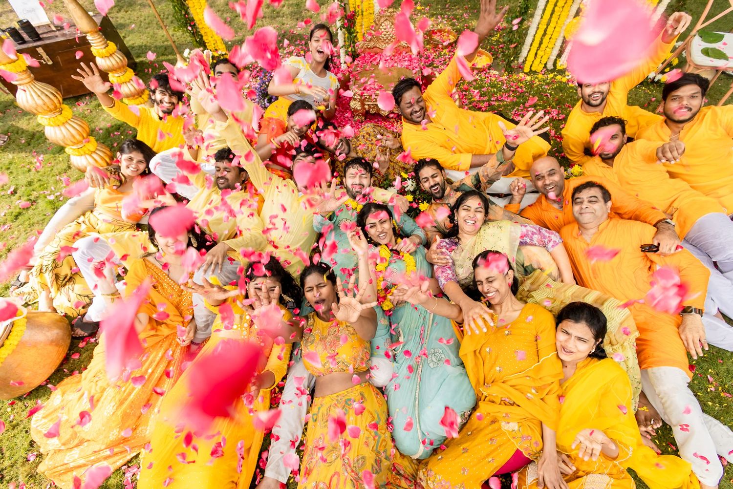 Aerial view of a joyful Indian Haldi ceremony group in yellow outfits tossing pink flower petals in the air.