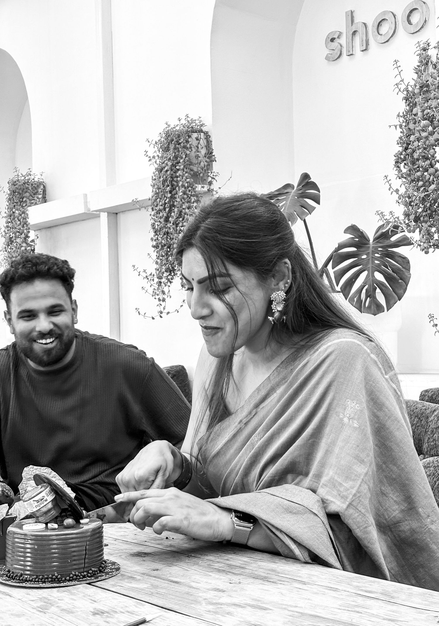 Woman cutting a cake at a cafe table while a smiling man watches in a black and white photo.