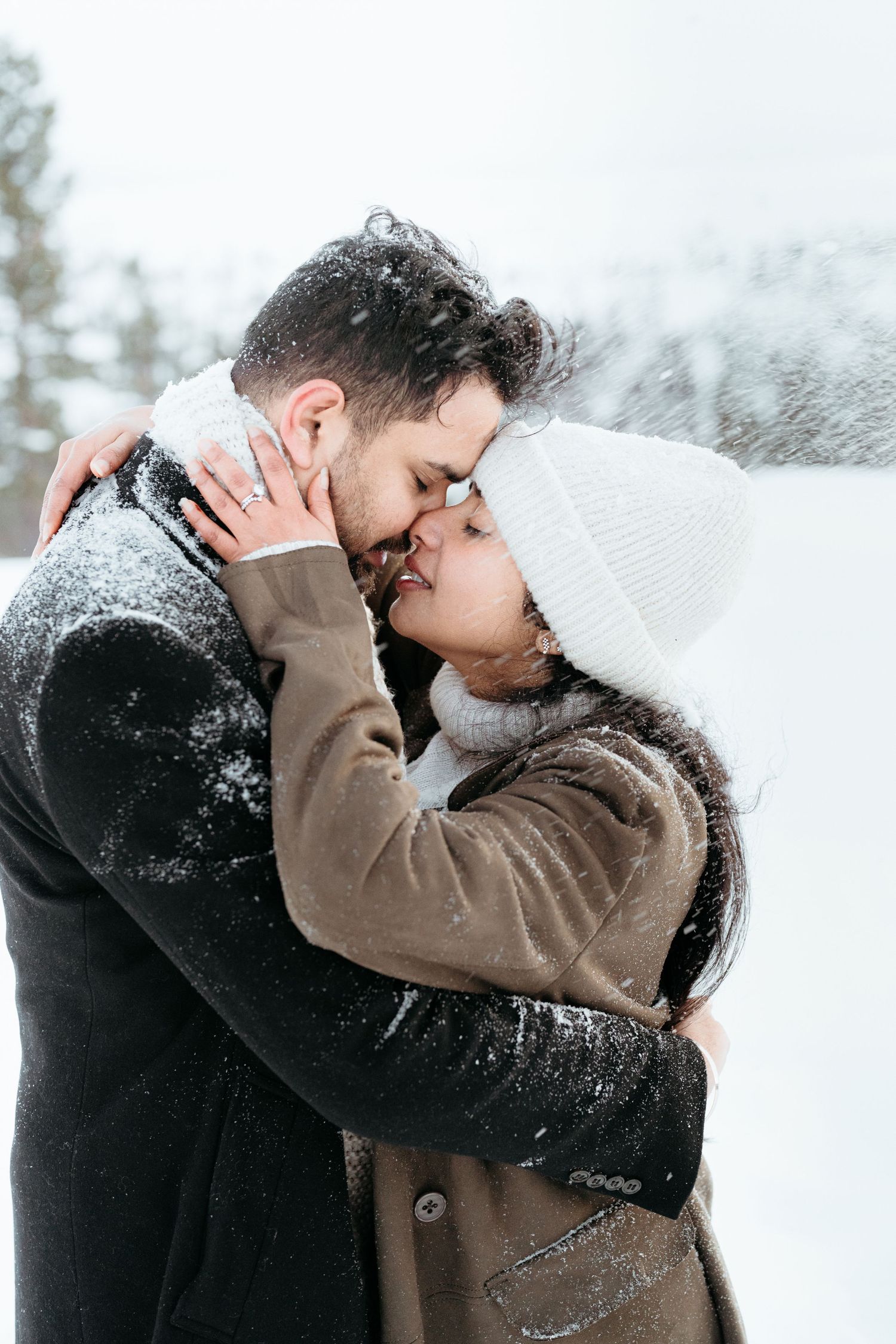Couple sharing a romantic kiss in snowy winter weather, wearing cozy coats and a white knit hat.