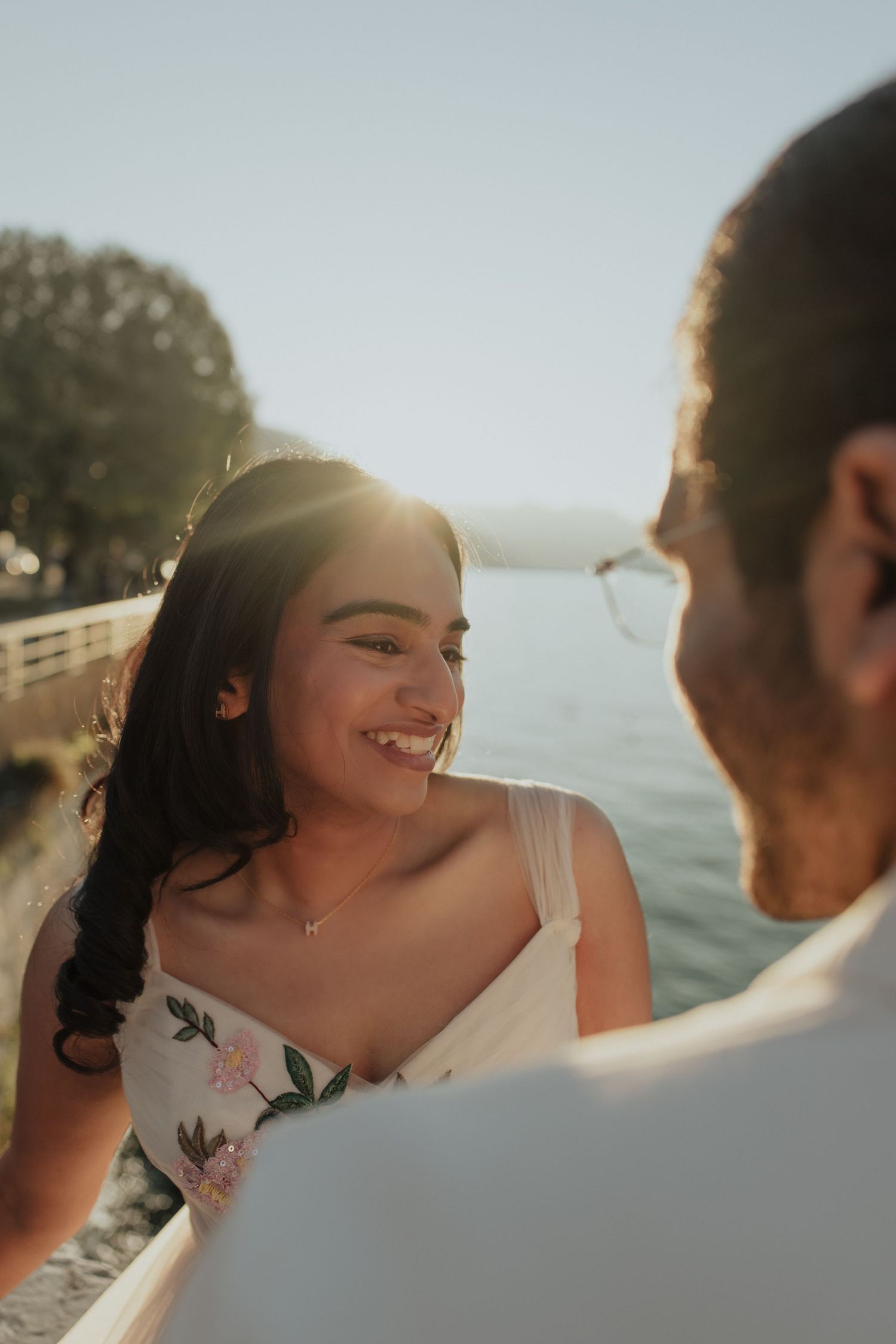 Smiling bride in floral dress gazing at groom with sunlit lake and mountains in background.
