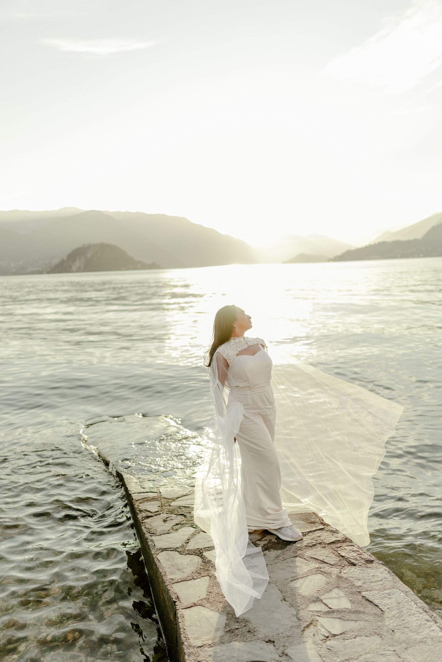 Bride in white dress stands on stone pier at sunset, veil flowing in wind over serene mountain lake.