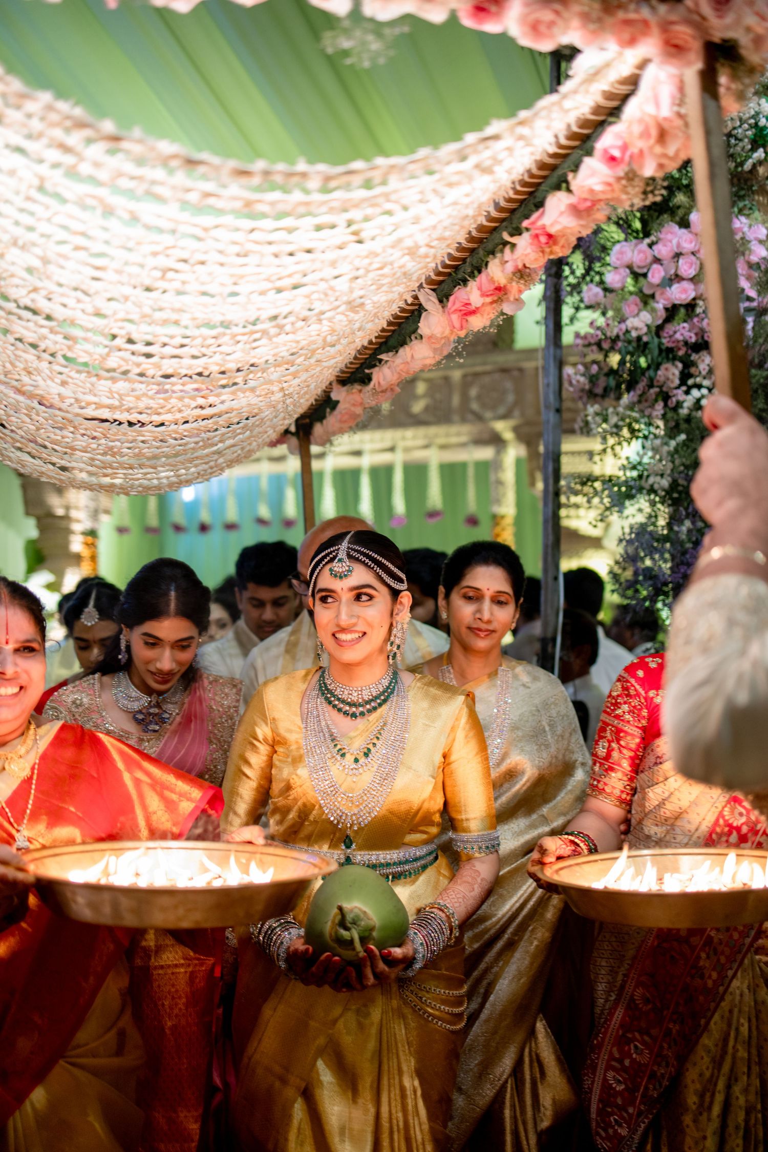 Radiant Indian bride in gold silk saree smiling while carrying ritual lamps in a decorated wedding mandap.