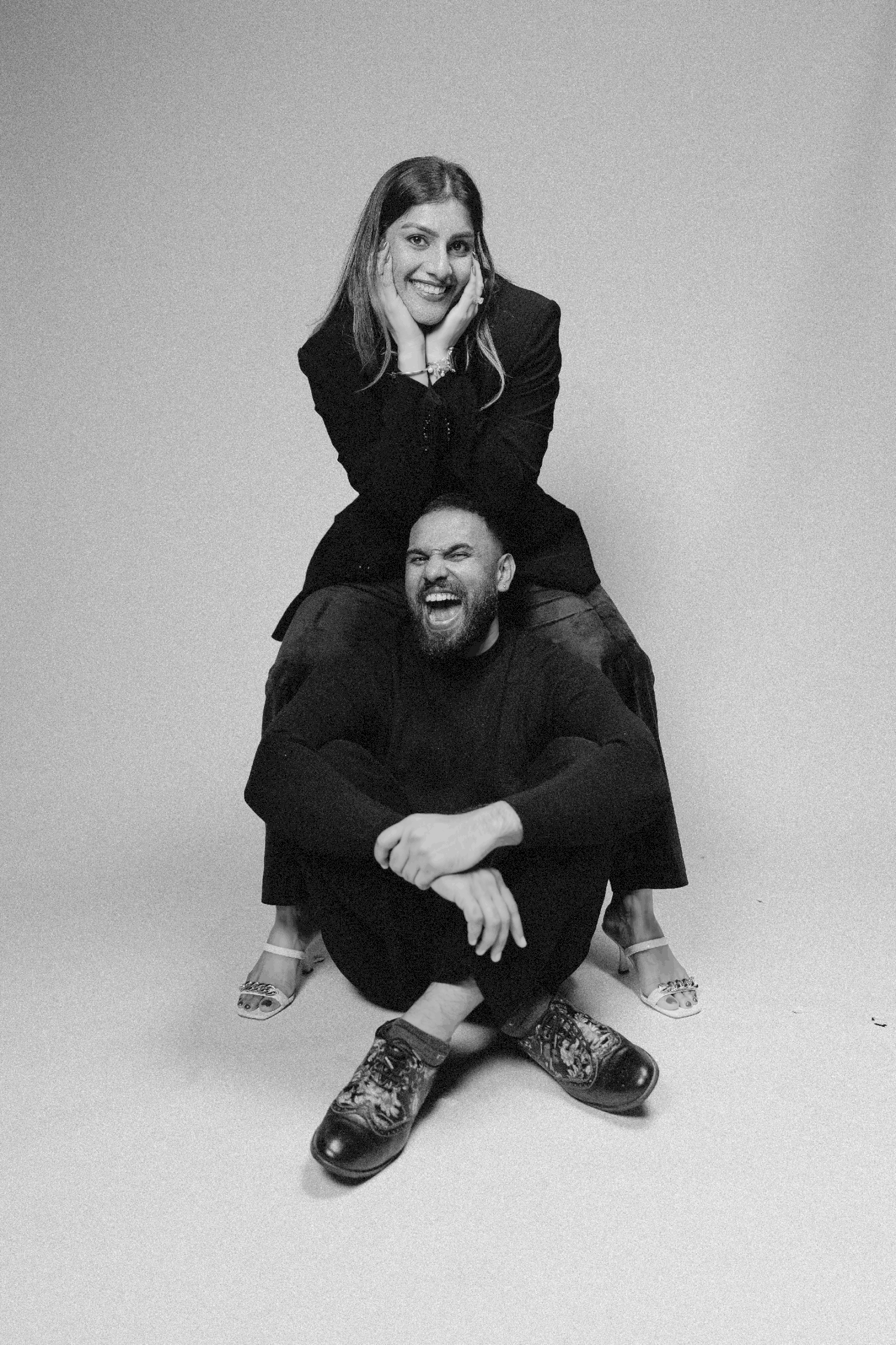 Black and white photo of a laughing couple in black outfits, woman sitting on mans shoulders in a studio.