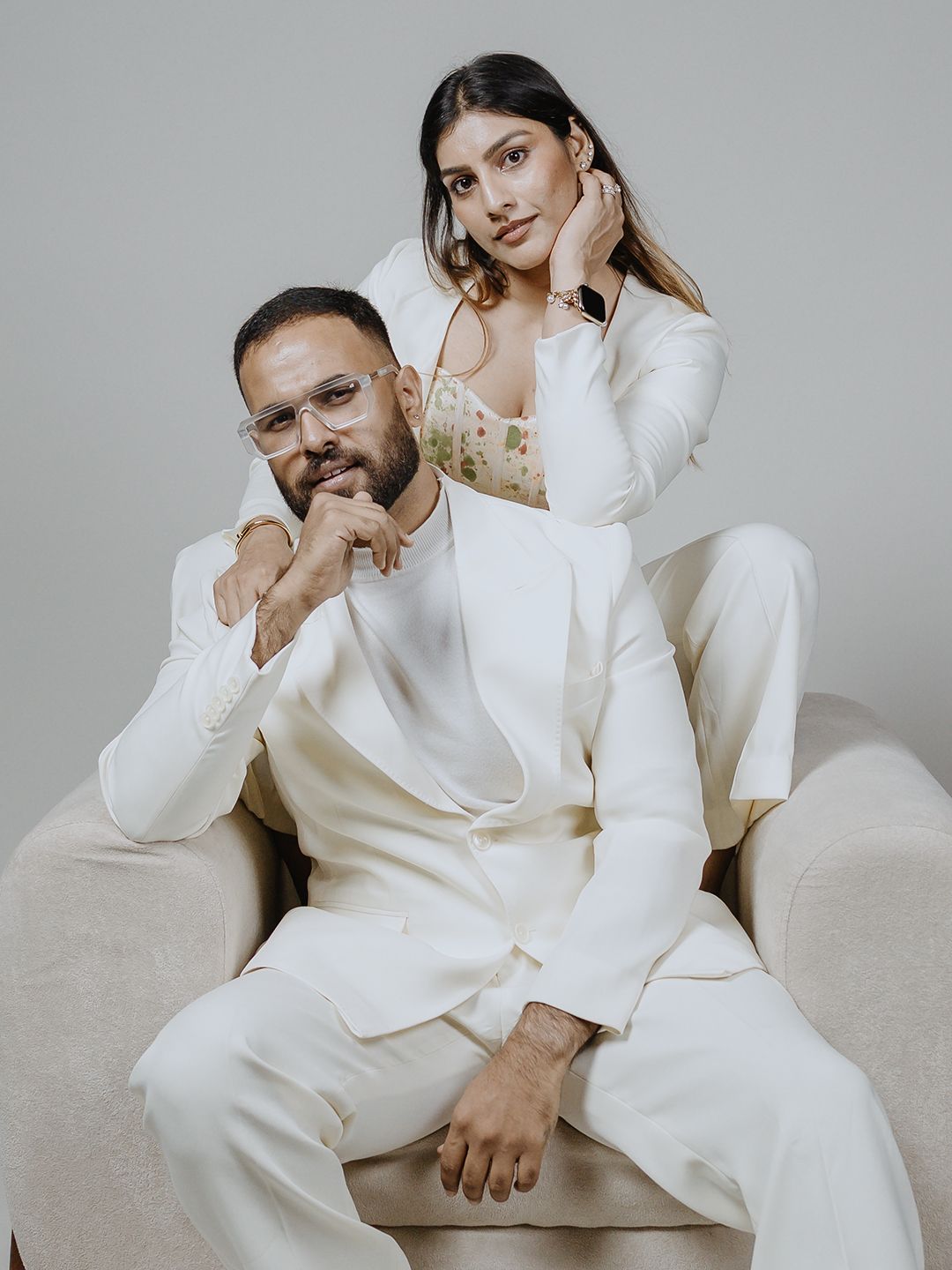 A man and woman in matching white suits pose elegantly in a minimalist studio setting.