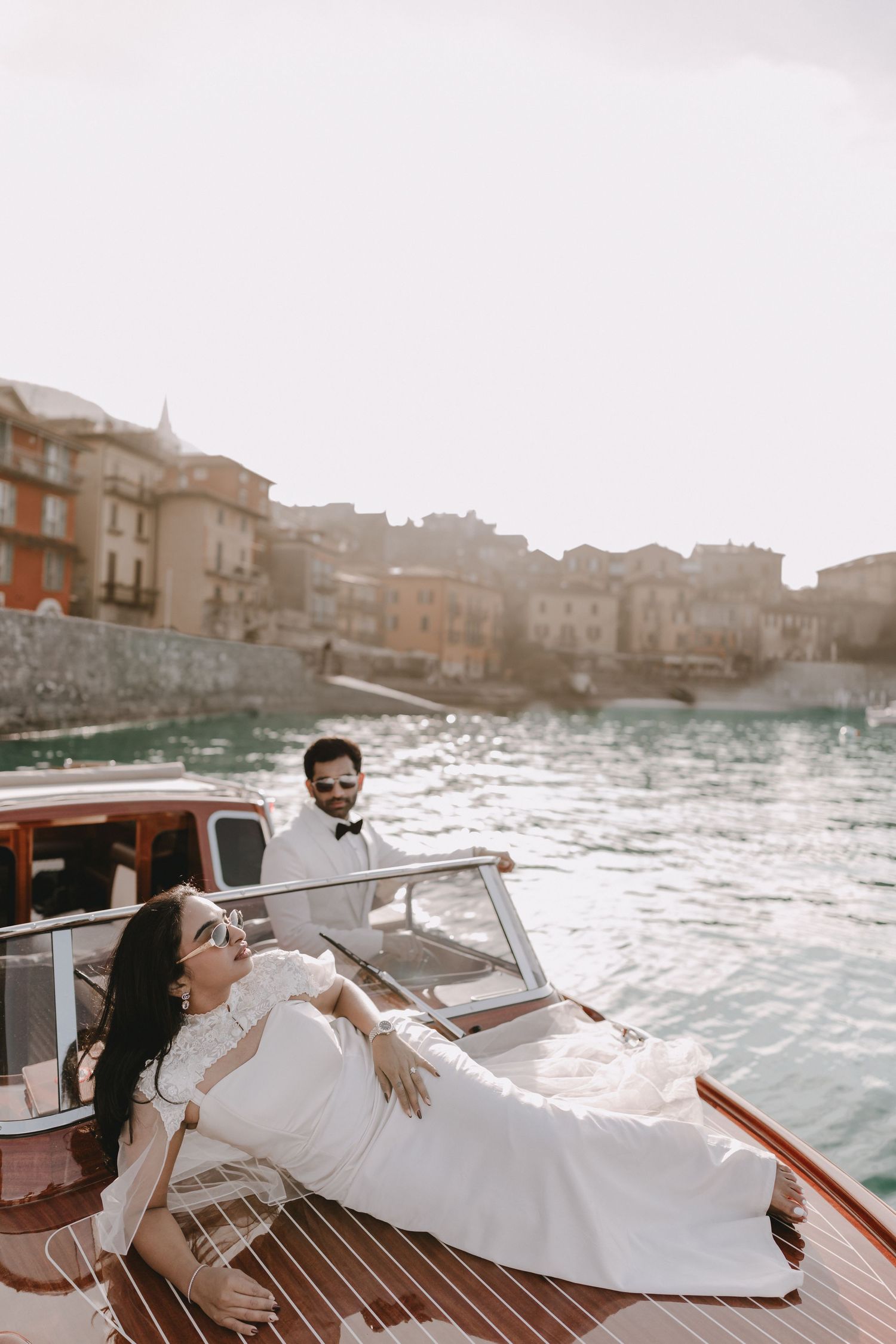 Bride in white gown reclines on luxury wooden boat while groom stands at helm, with Italian lakeside village backdrop.