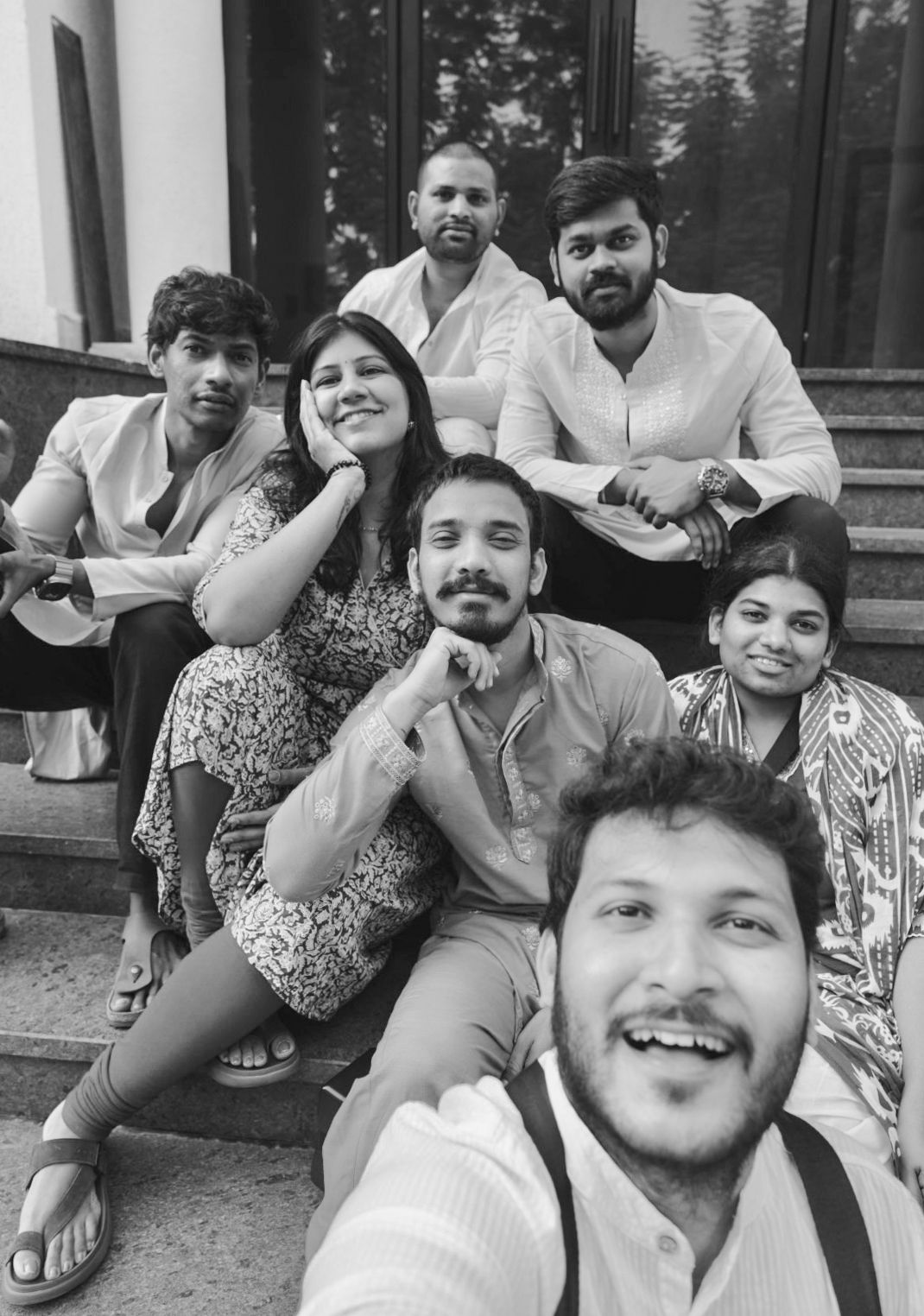 Group of seven friends posing together on outdoor steps for a black and white selfie photo.