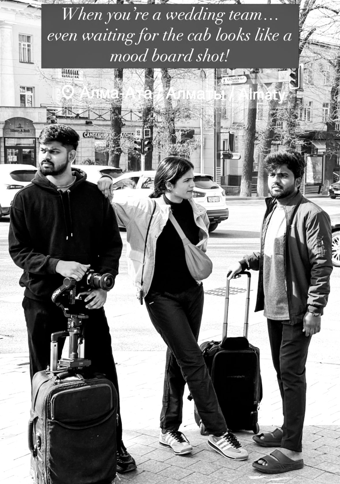Black and white photo of three wedding photographers waiting for a cab with luggage and camera equipment on a city street.