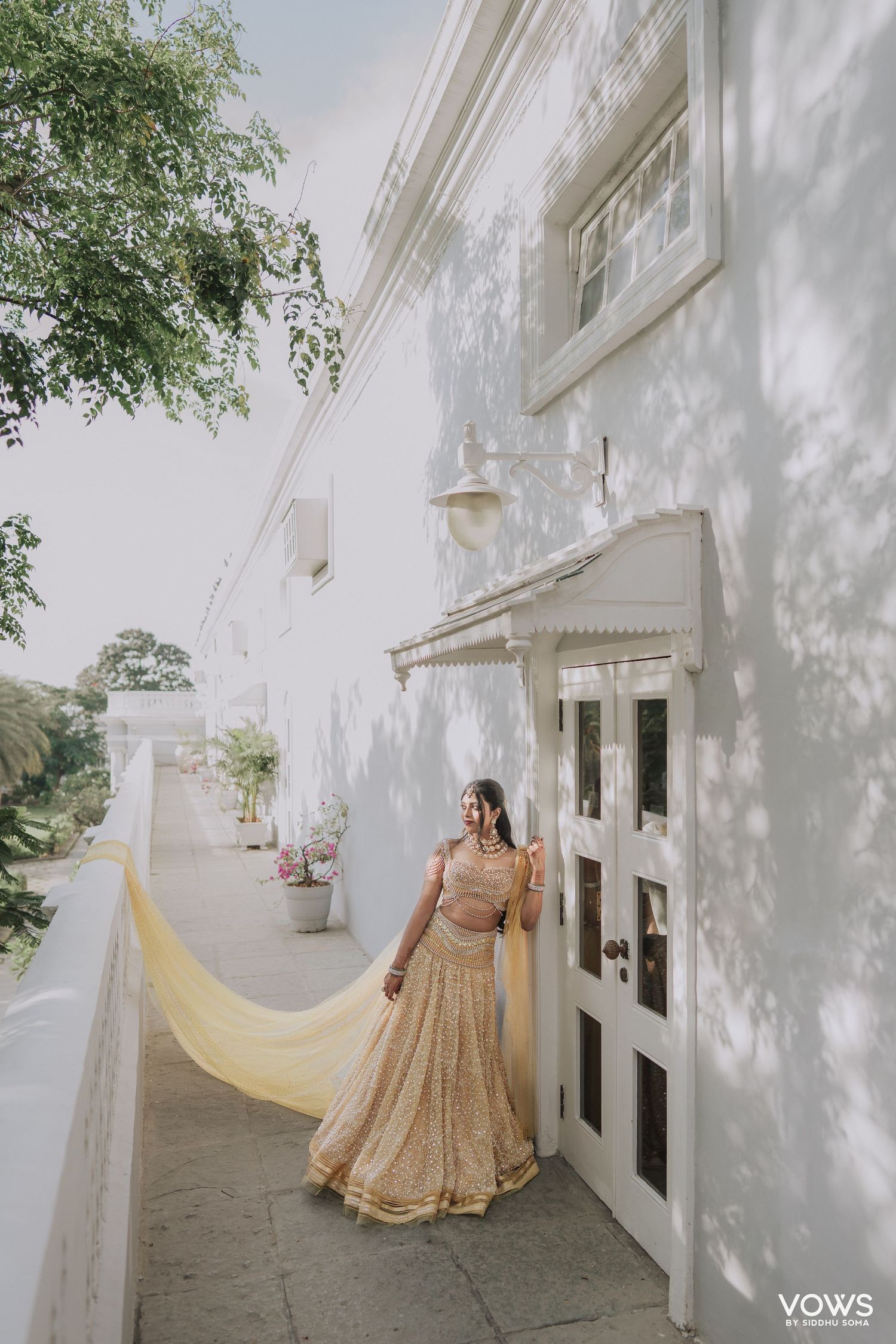 Bride in golden lehenga with flowing yellow dupatta poses along white colonial building corridor.