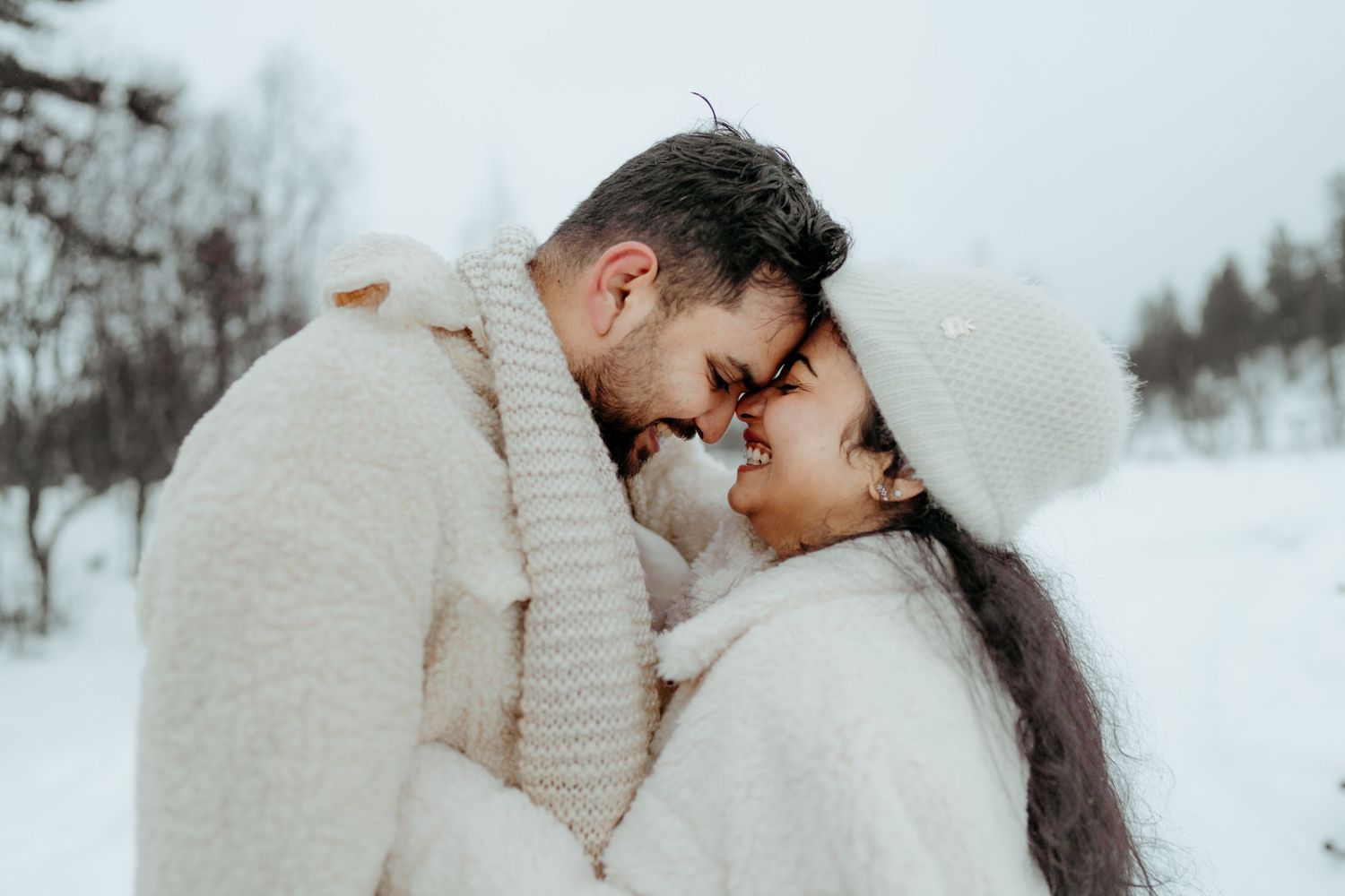Romantic couple in white winter outfits touching foreheads in snowy landscape, sharing a tender moment outdoors.
