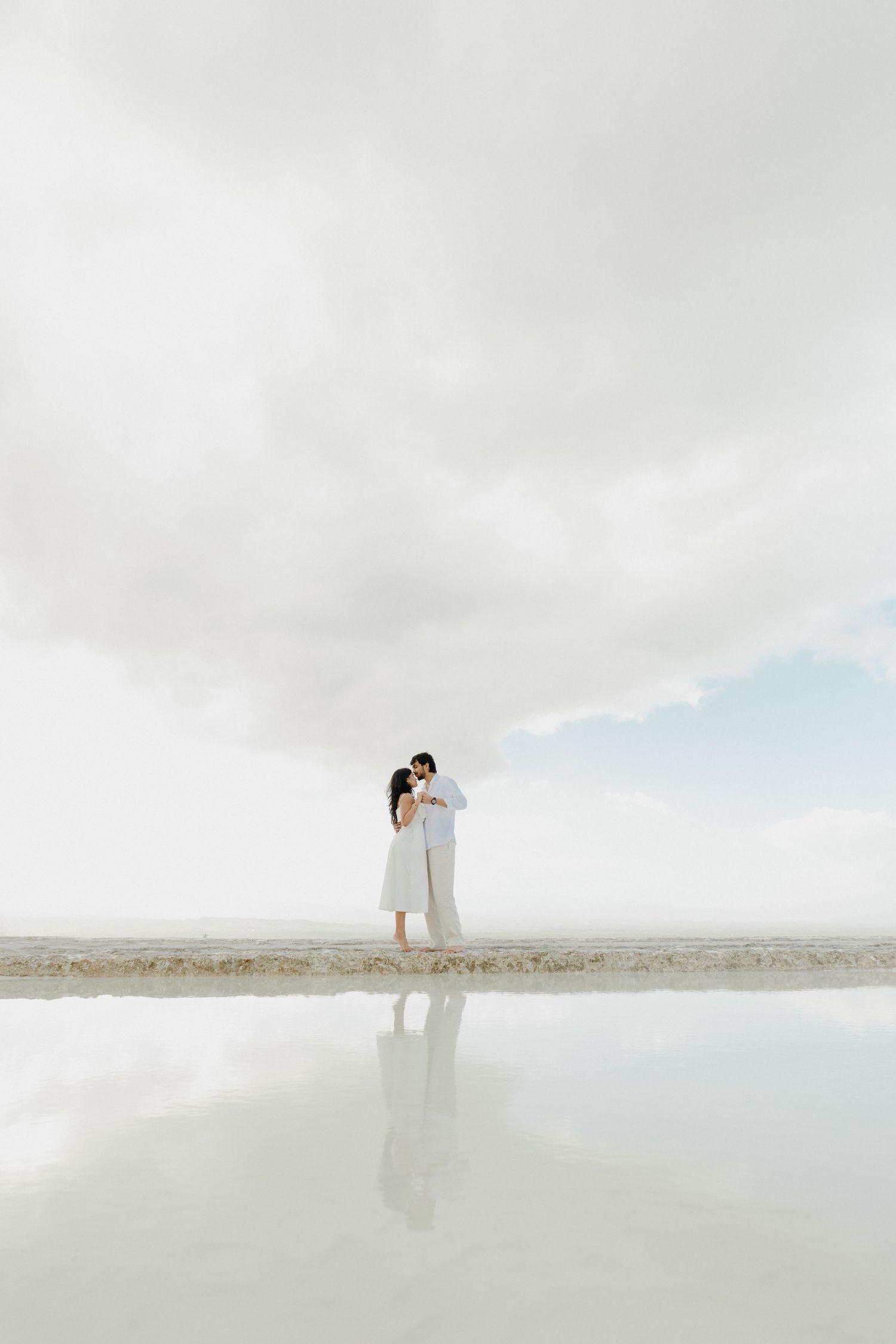 Couple embracing and kissing by a reflective salt flat under dramatic cloudy skies.