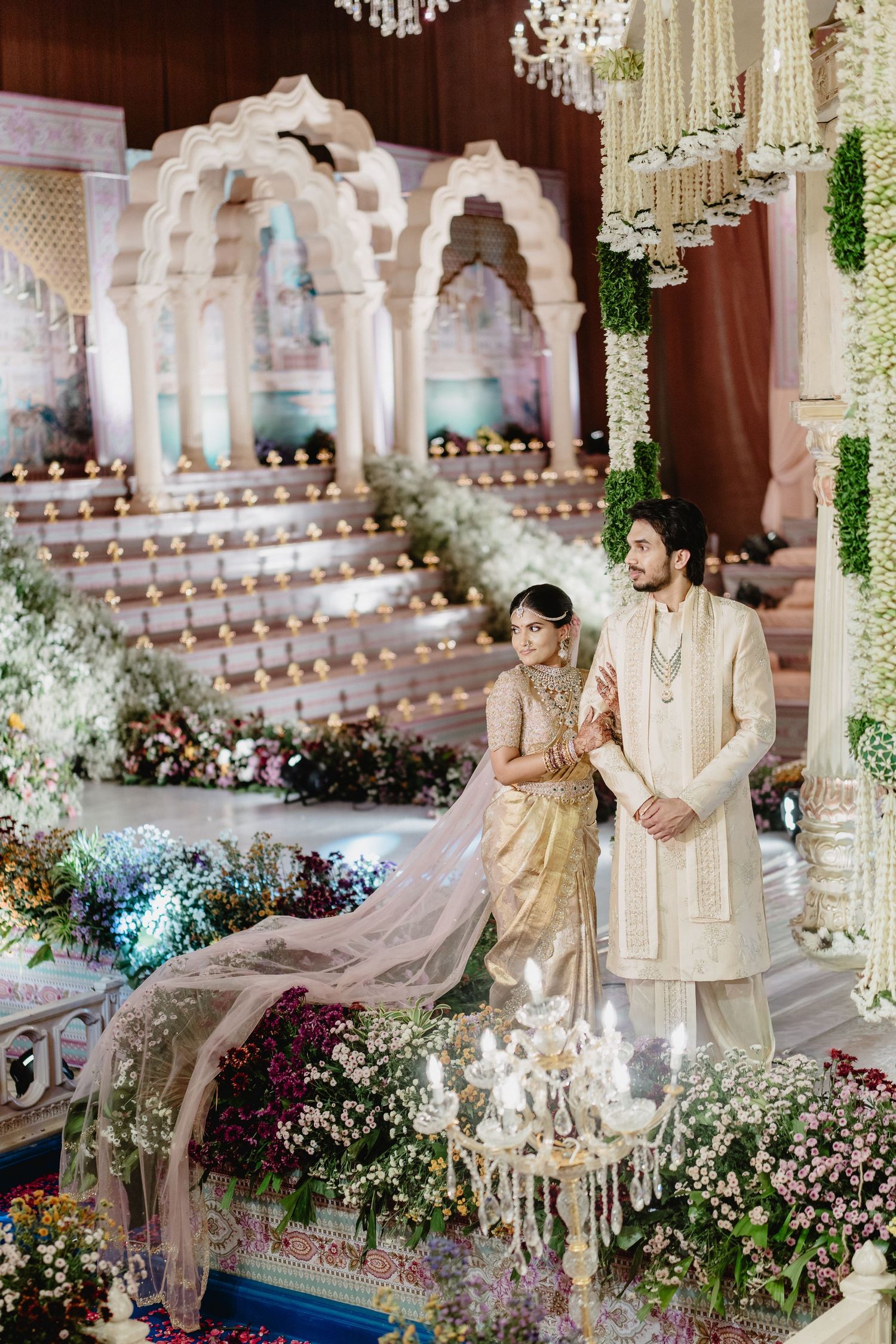 Indian bride and groom in gold attire pose at ornate floral wedding mandap with white arch backdrop and chandelier.