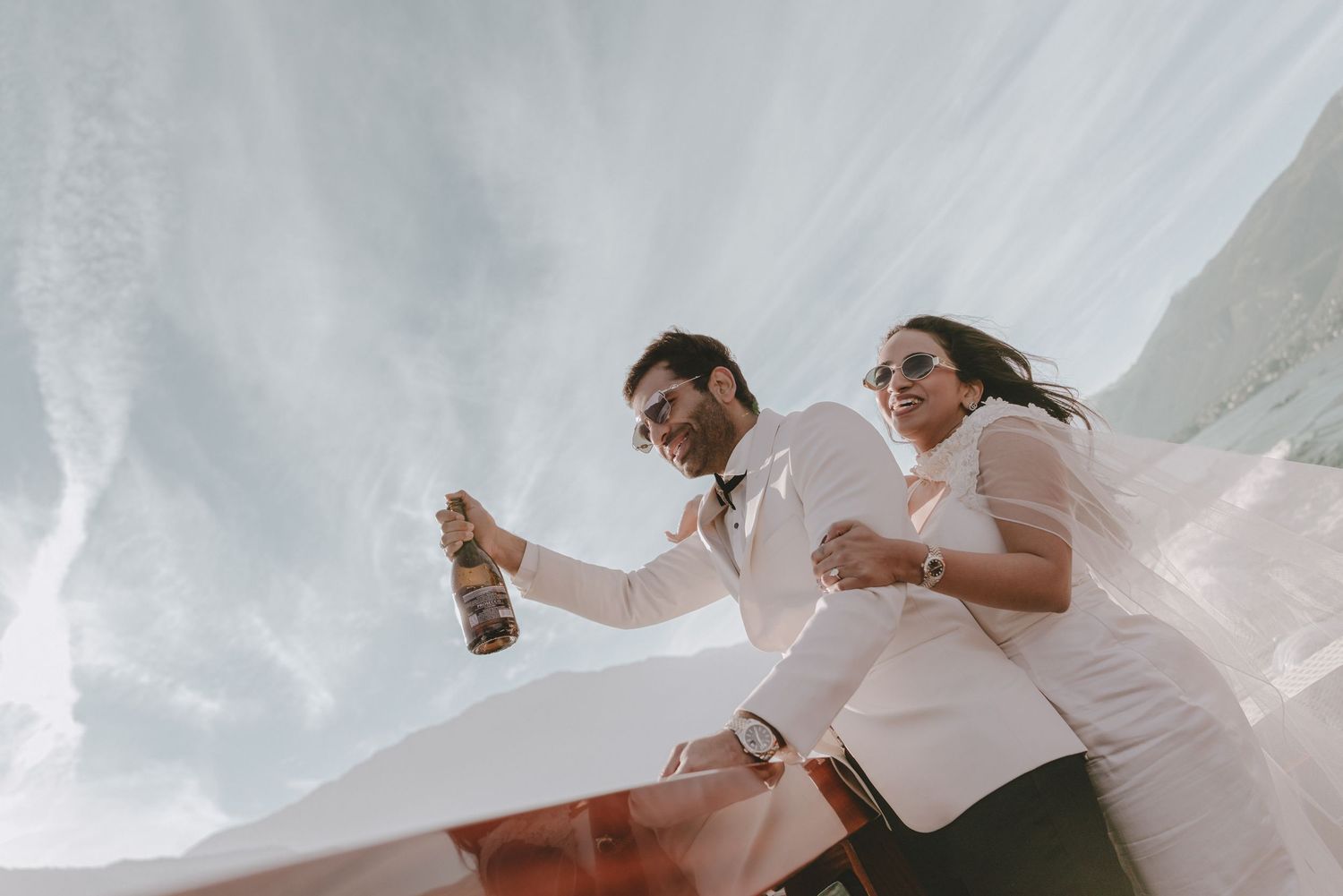 Couple in white attire celebrating outdoors, man pouring champagne bottle against dramatic cloudy sky.