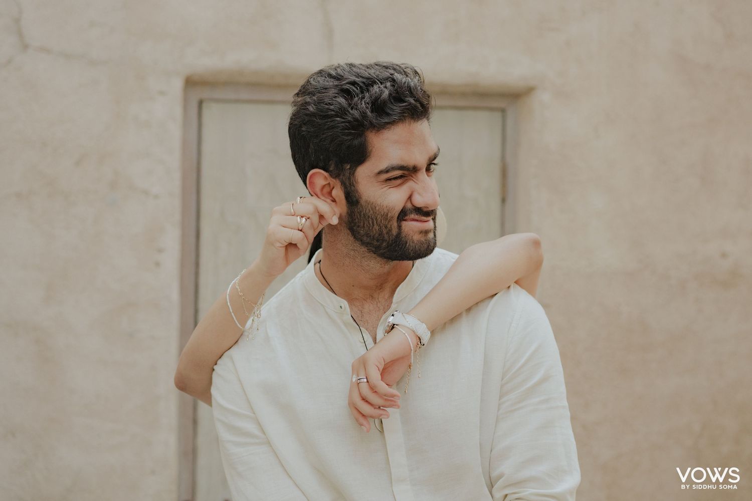 Young man in white linen shirt adjusting earring outdoors against a beige stone wall.