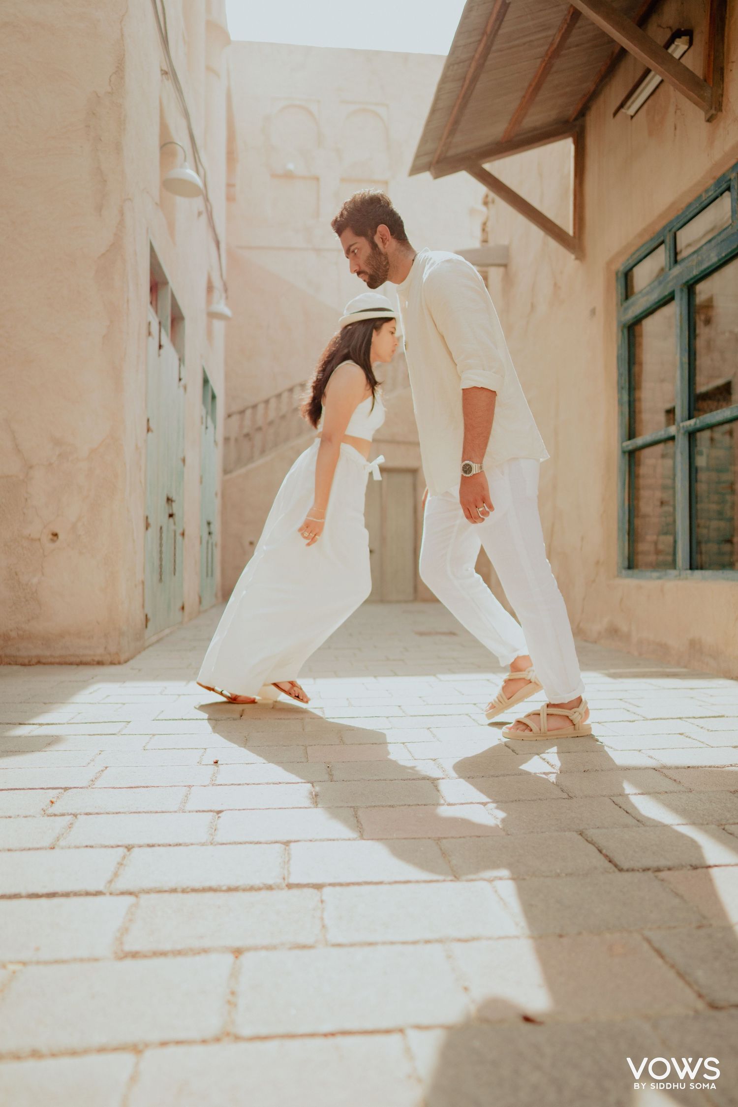 Couple in white outfits walking together on stone courtyard in sunny outdoor setting for a wedding shoot.