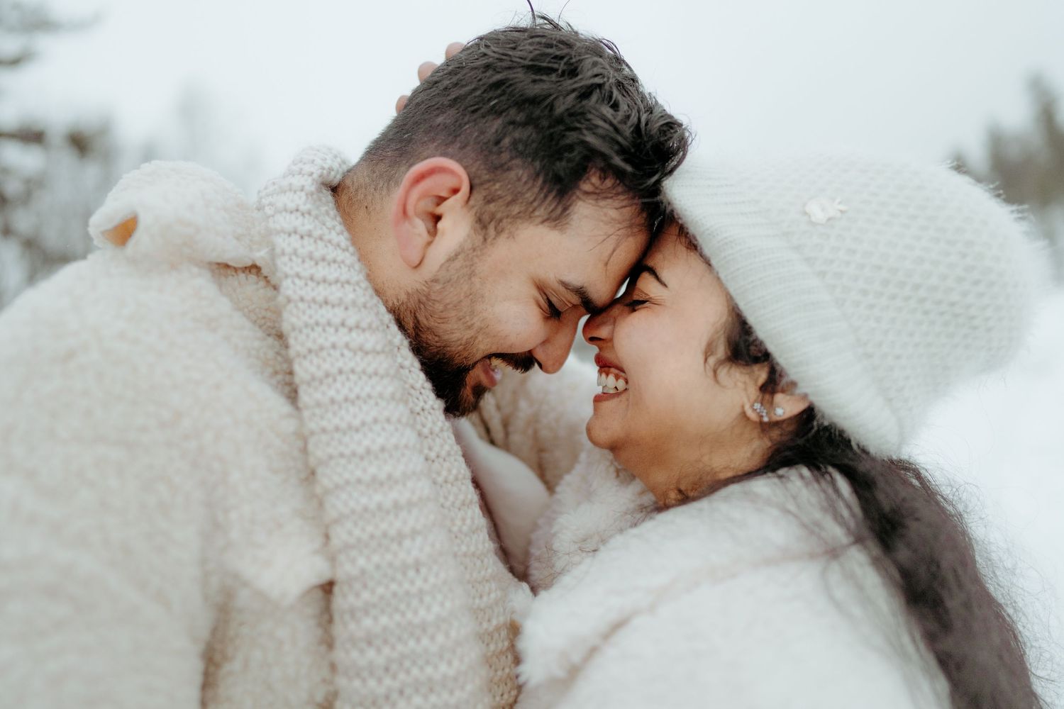 Romantic couple in white winter outfits touching foreheads and smiling in snowy outdoor setting.