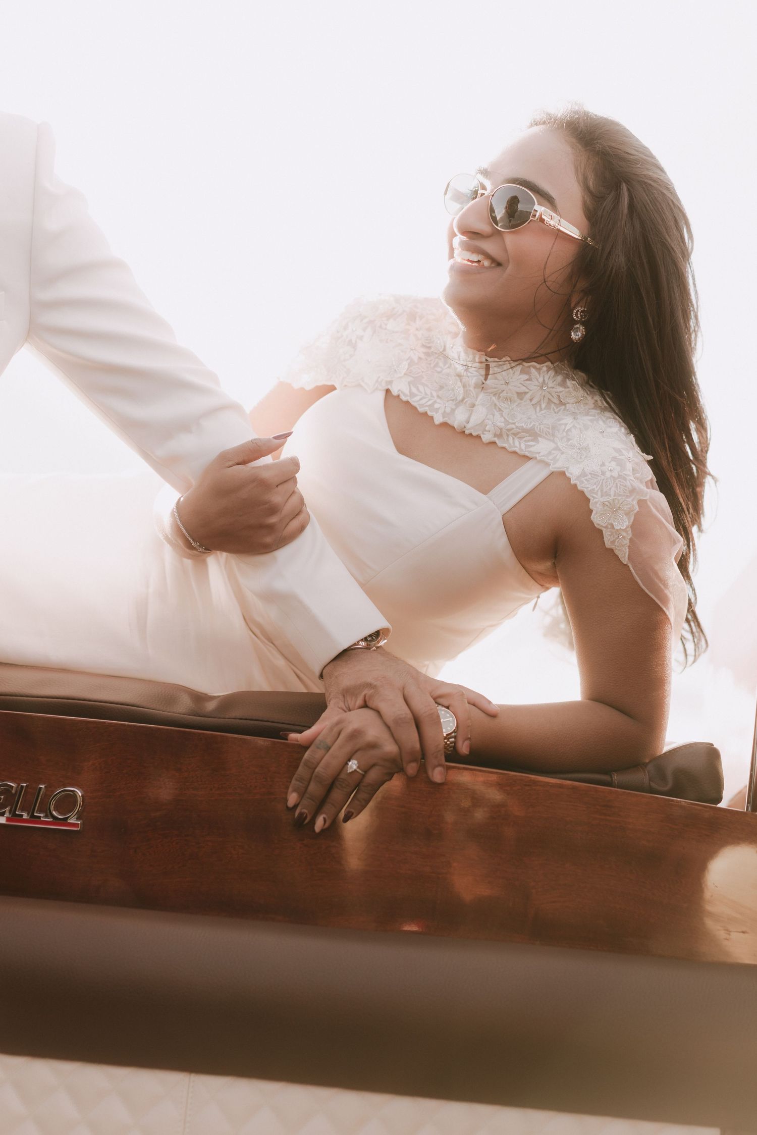 Radiant bride in white lace dress and sunglasses smiles joyfully while seated in a luxury wooden boat.