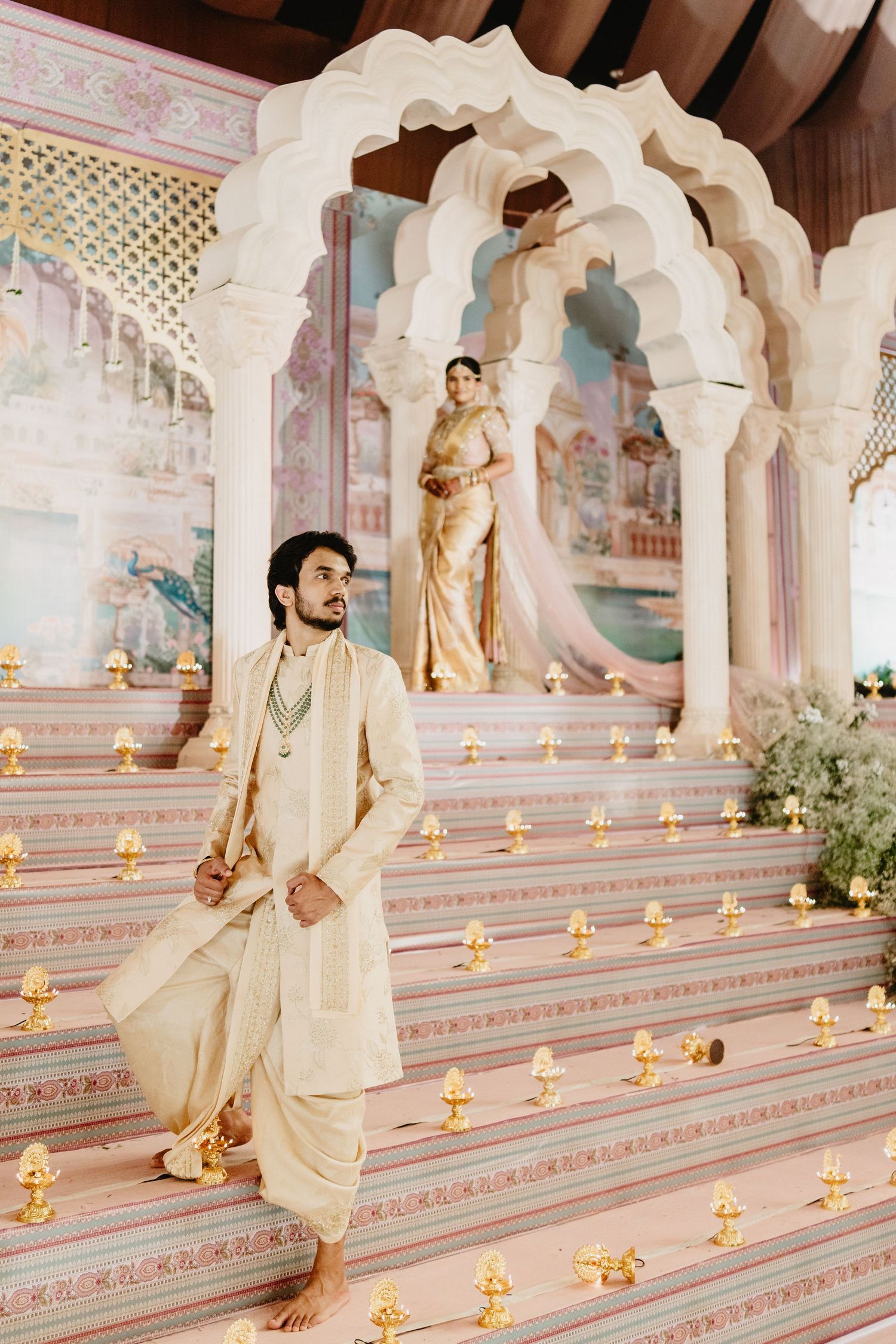 Groom in cream sherwani descending ornate temple steps with bride standing in golden archway above.