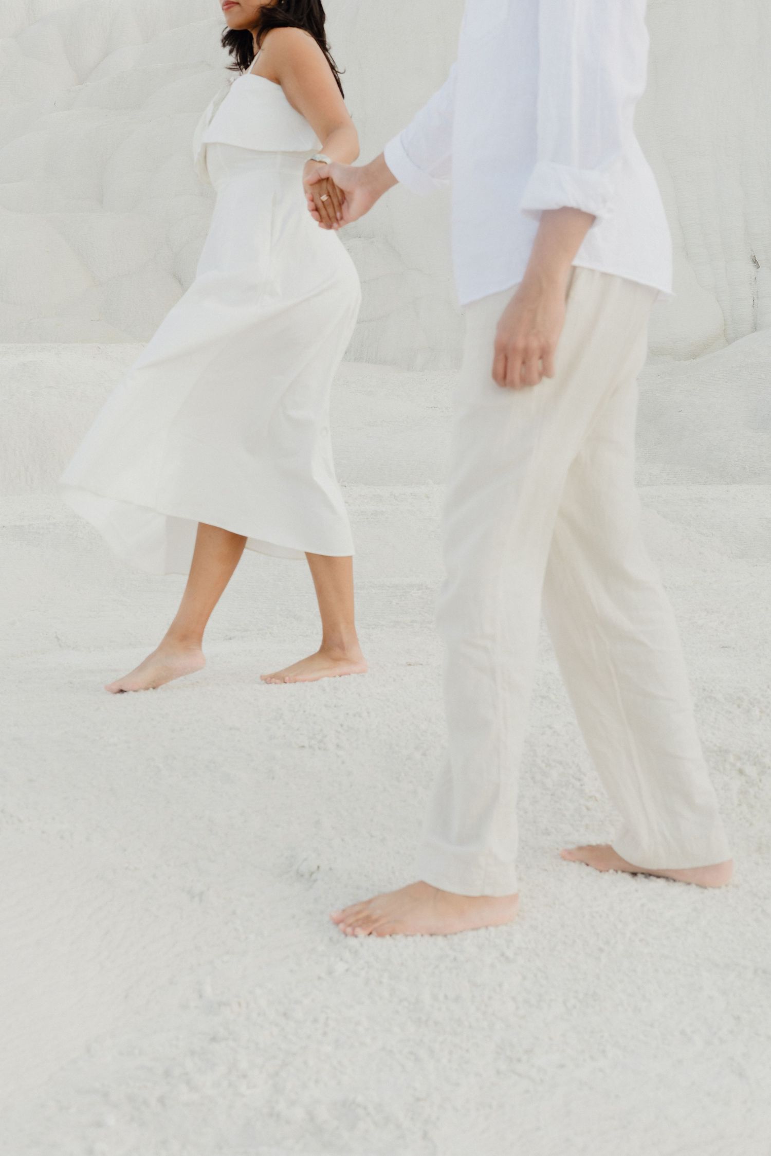 Barefoot couple in white outfits hold hands while walking on bright white travertine terraces at Pamukkale.