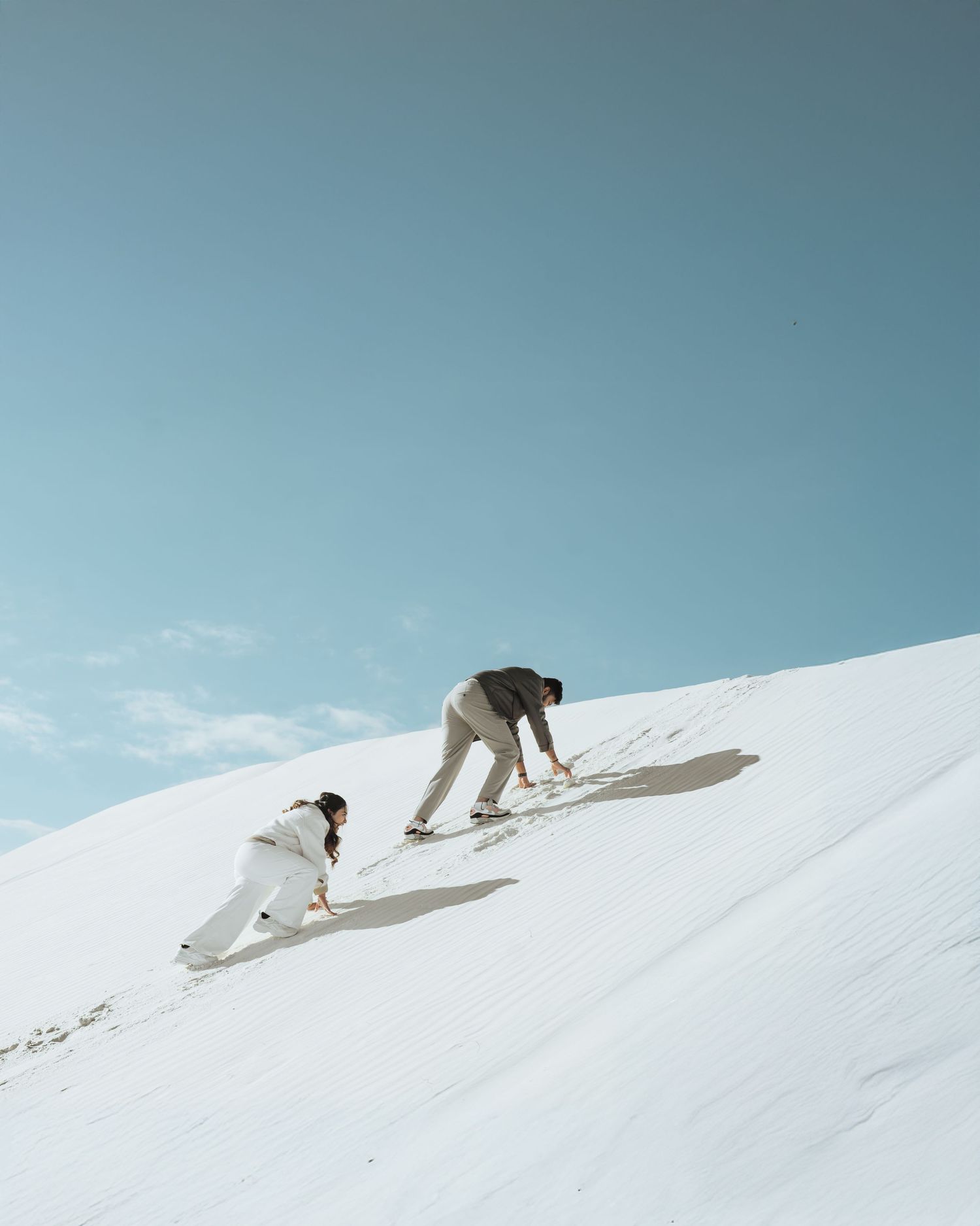 Two people climbing a bright white sand dune under a clear blue sky, casting long shadows on the slope.