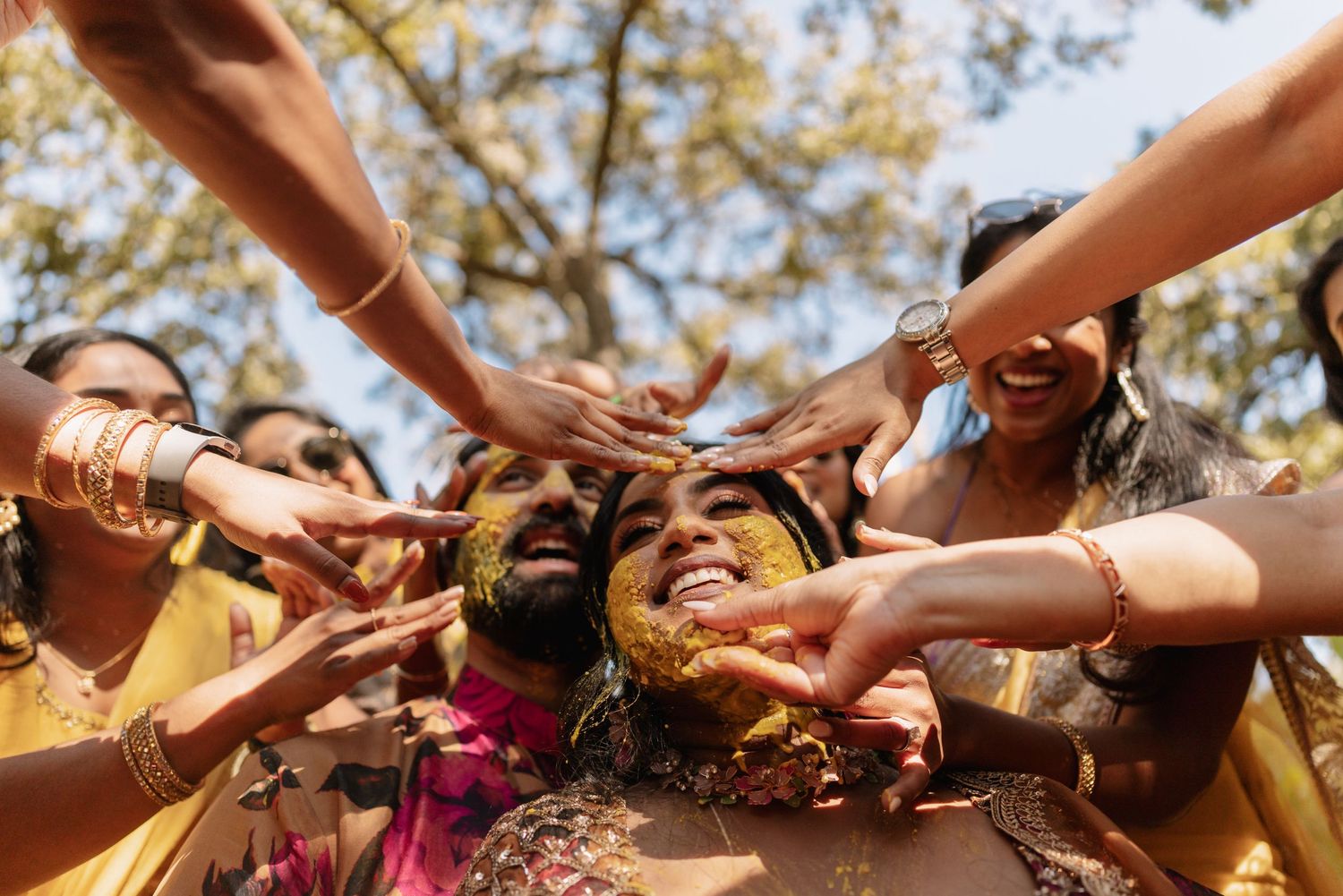 Group of friends joyfully applying haldi paste on a laughing person during a traditional Indian wedding ceremony outdoors.