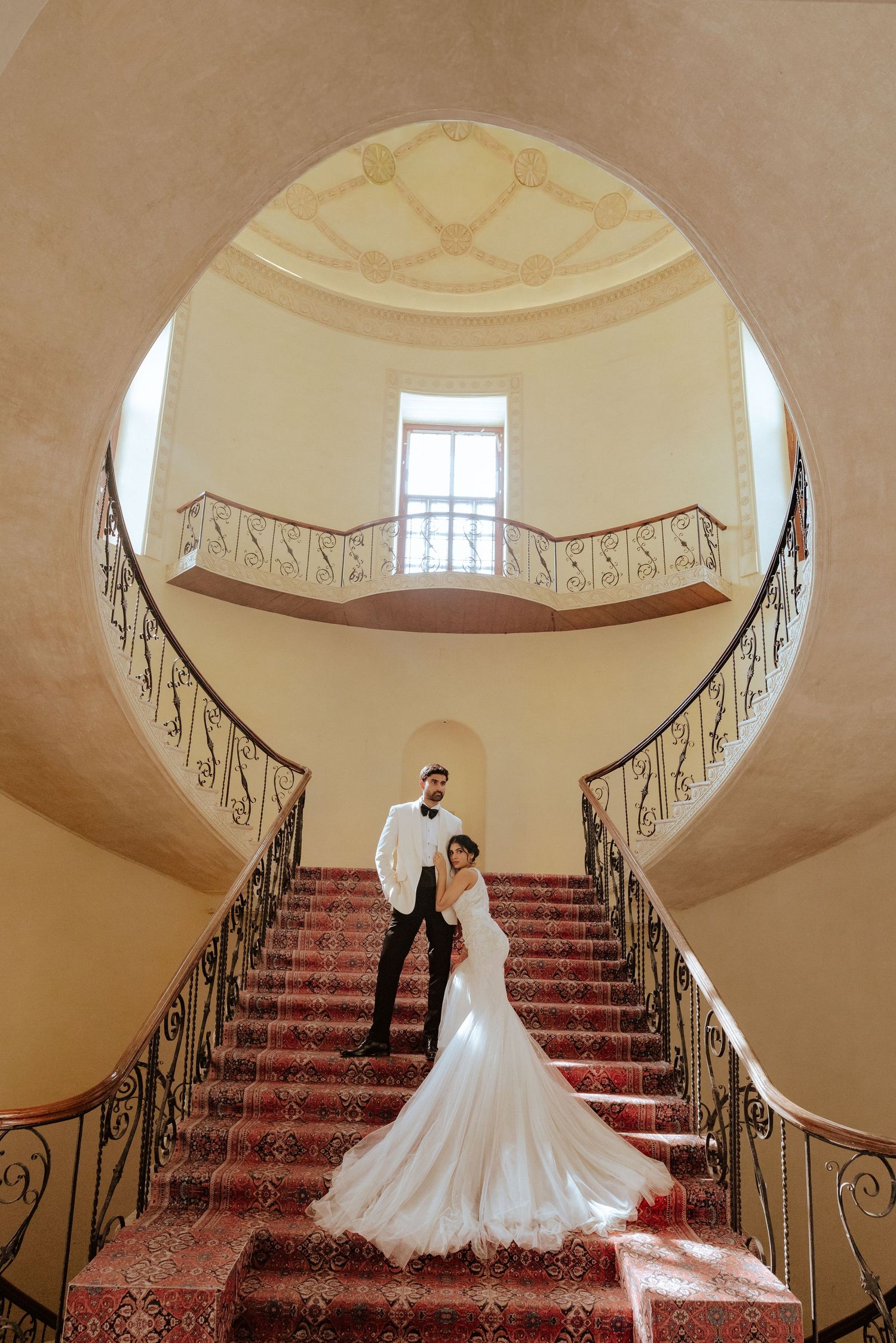 Bride and groom posing on elegant red-carpeted spiral staircase in a grand venue with ornate balcony.