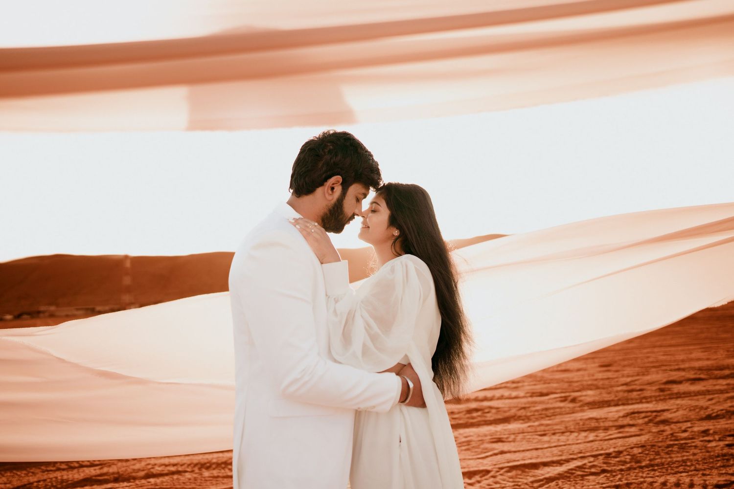 Couple in white outfits embrace in a desert at sunset as flowing peach fabric billows around them in a romantic pre-wedding photoshoot.