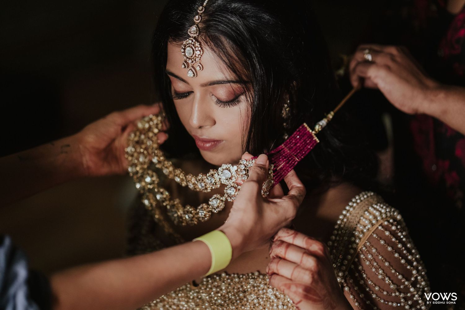 Indian bride in gold lehenga getting jewelry fastened during bridal preparation ceremony.